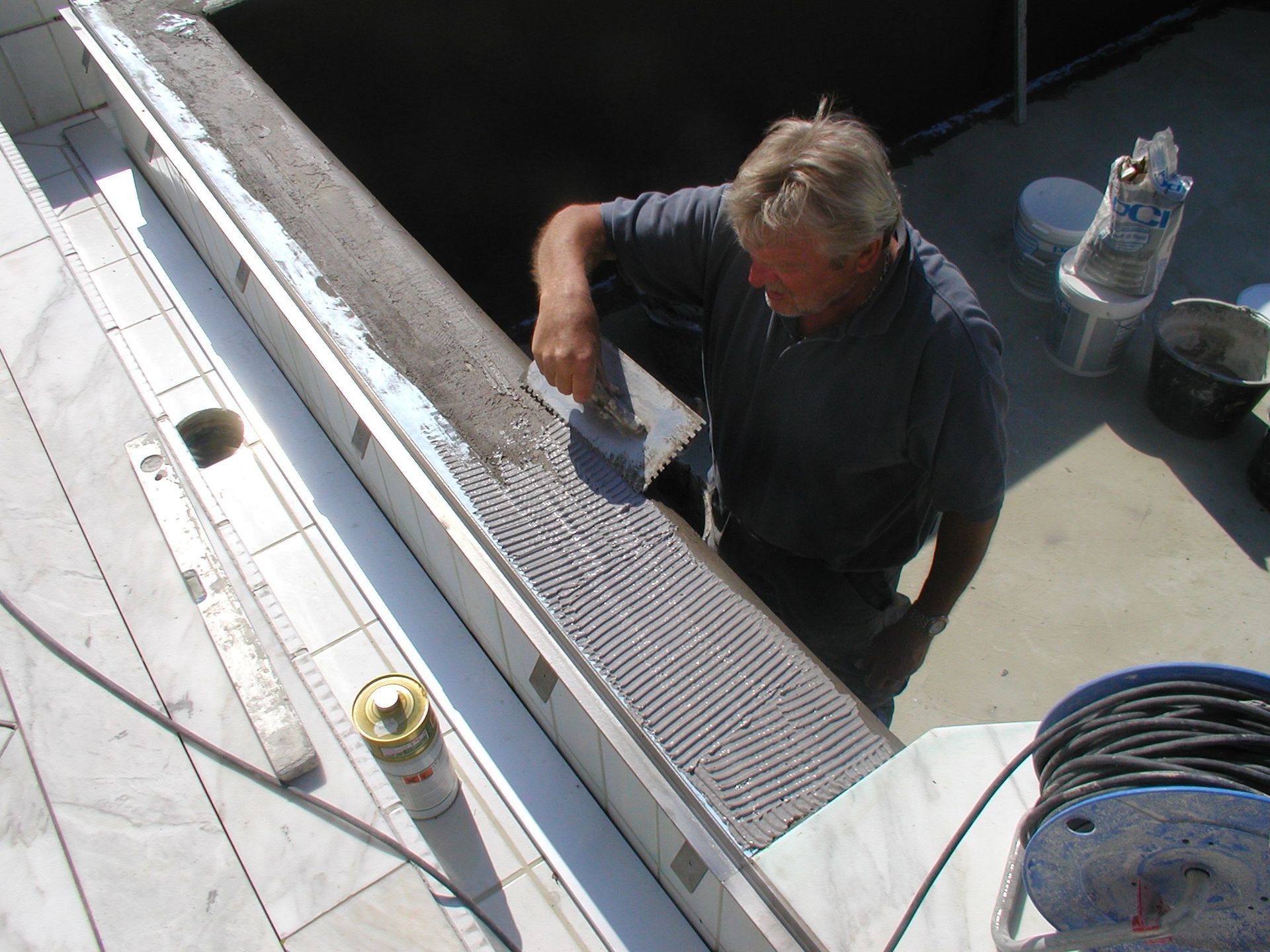 Mann trägt mit einer Kelle Klebstoff auf einen Sims auf und bereitet ihn für das Fliesenlegen vor. Im Freien.