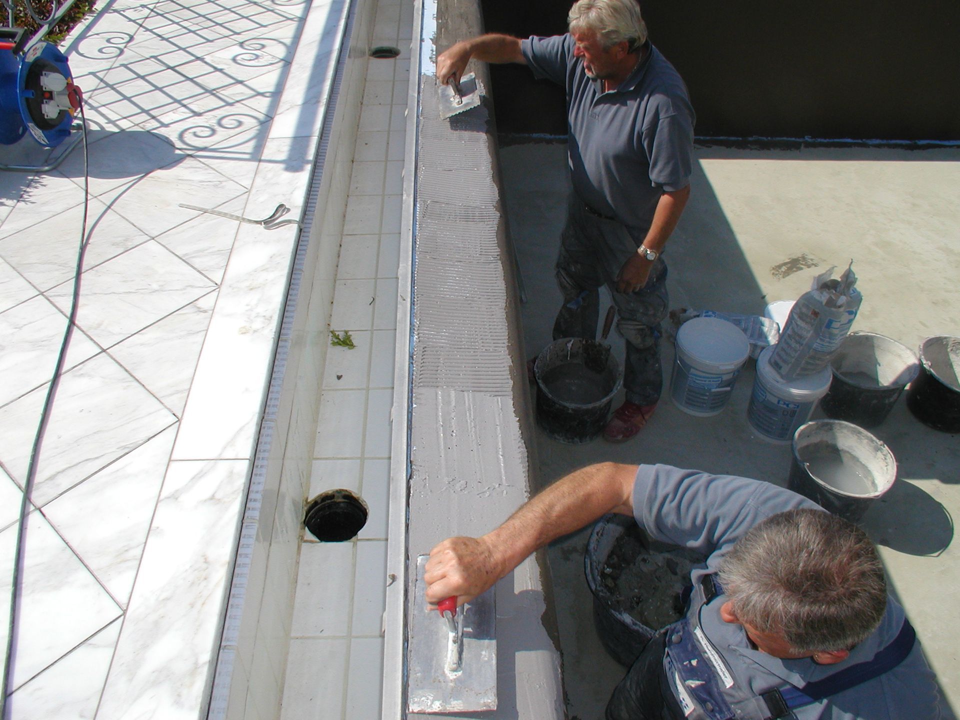 Zwei Männer fliesen eine lange Dachrinne und tragen im Freien mit Kellen grauen Mörtel auf.