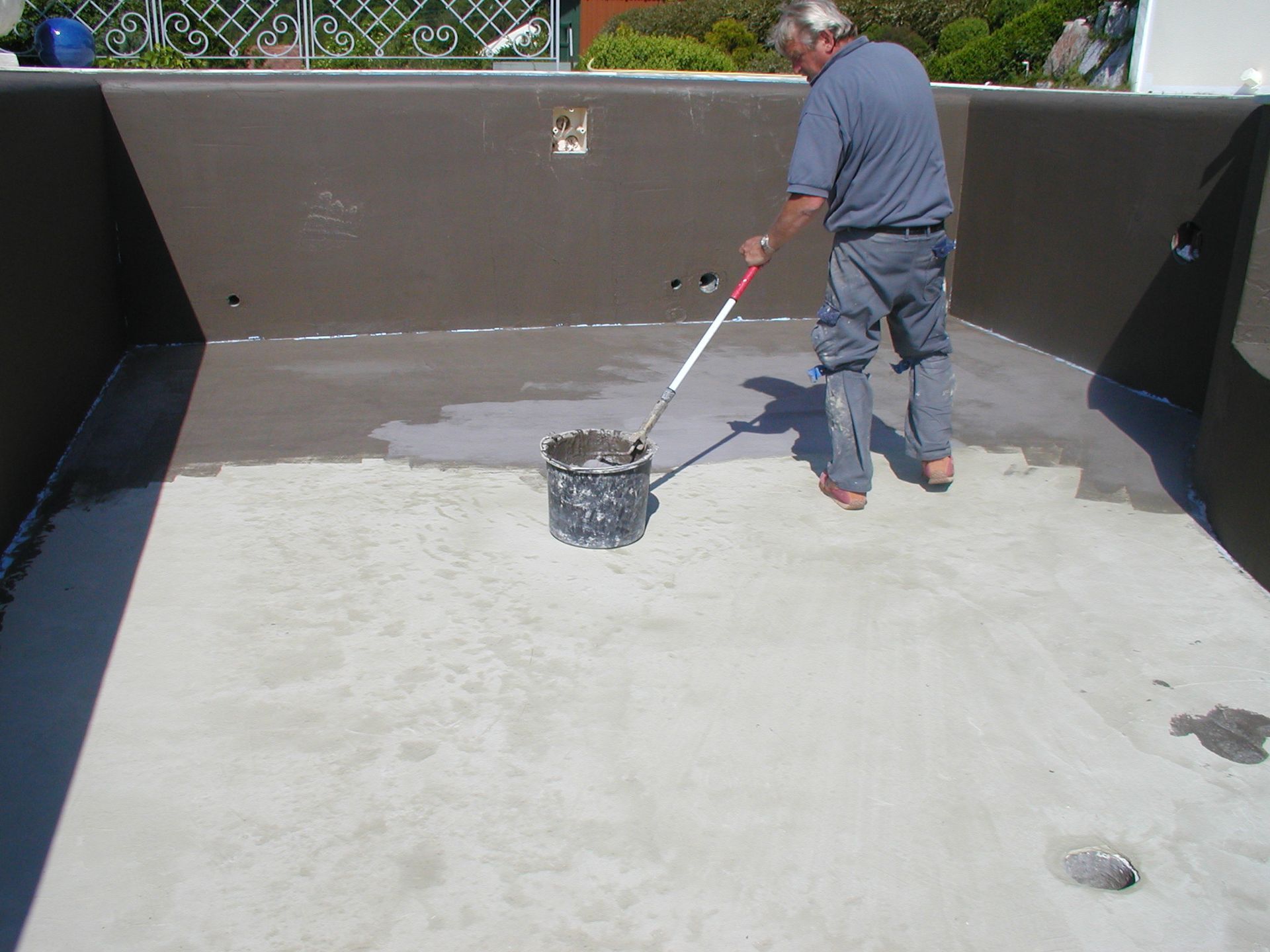 Ein Mann streicht mit einer Rolle die Innenseite eines dunklen Pools in einem Betonbecken.