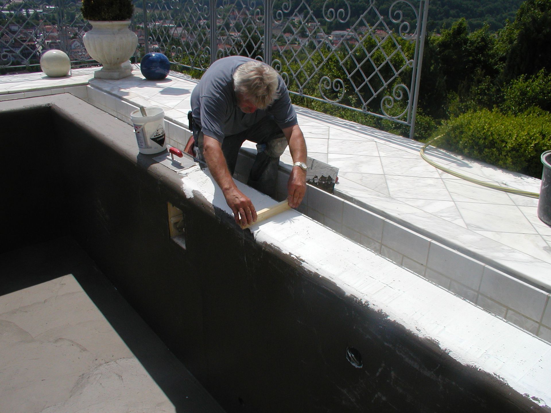 Mann fliest den Beckenrand. Er kniet und verteilt mit einem Werkzeug weißen Mörtel, im Außenbereich, sonnig, in der Nähe eines weißen Geländers.