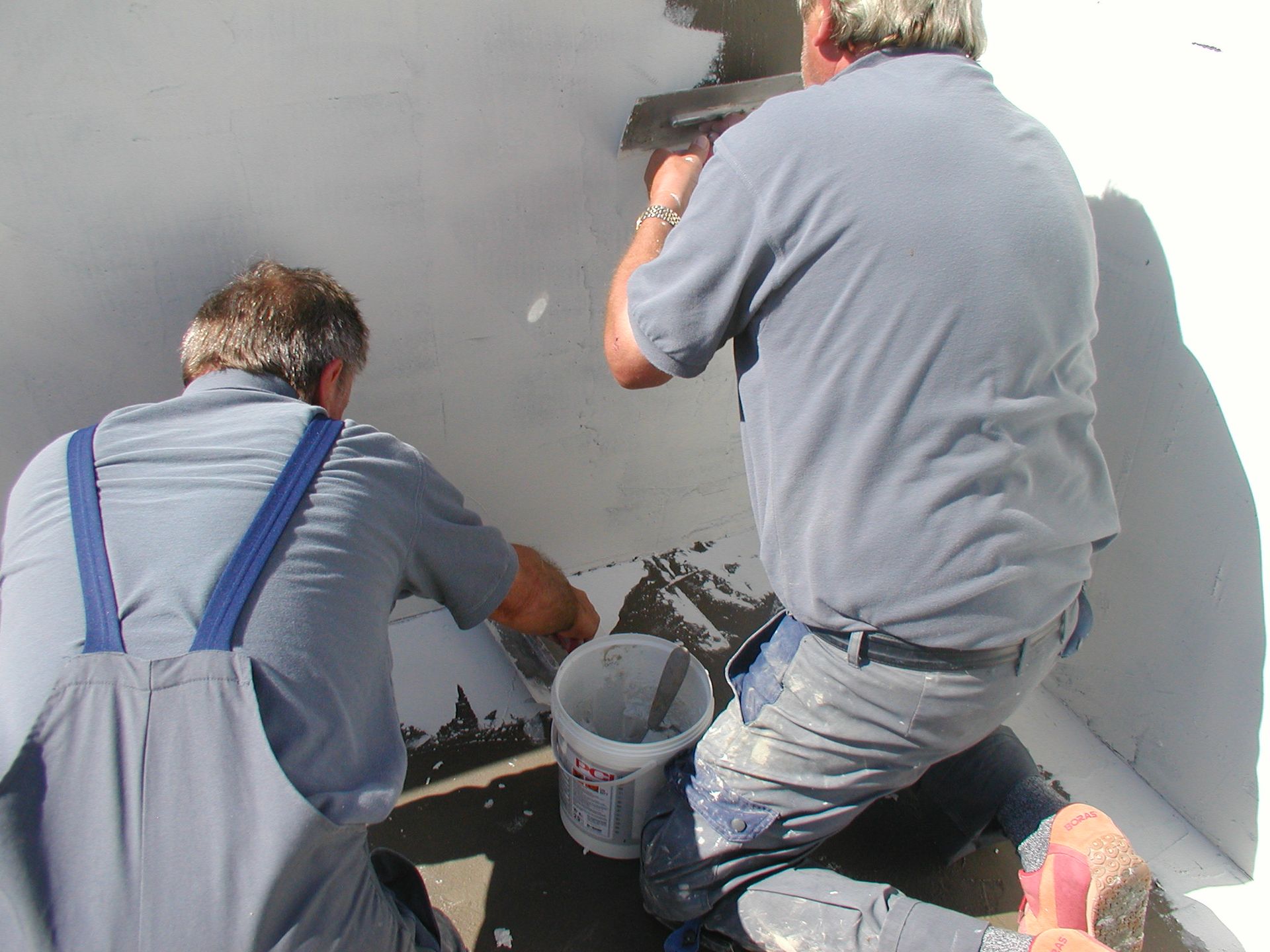 Zwei Männer flicken eine weiße Decke mit Werkzeug und einem Eimer Material.