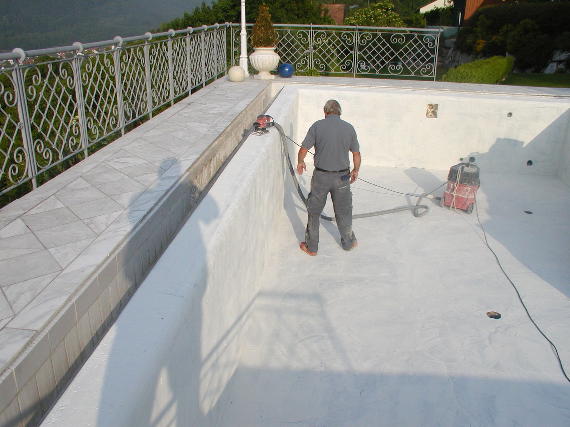 Mann arbeitet mit Werkzeugen an einem leeren weißen Pool. Steinterrasse und Zaun im Hintergrund.