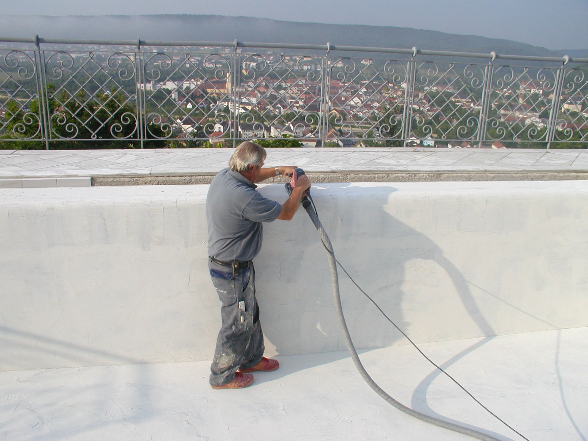 Mann benutzt ein Werkzeug an einer weißen Wand im Freien mit Blick auf eine Stadt mit Metallgeländer.