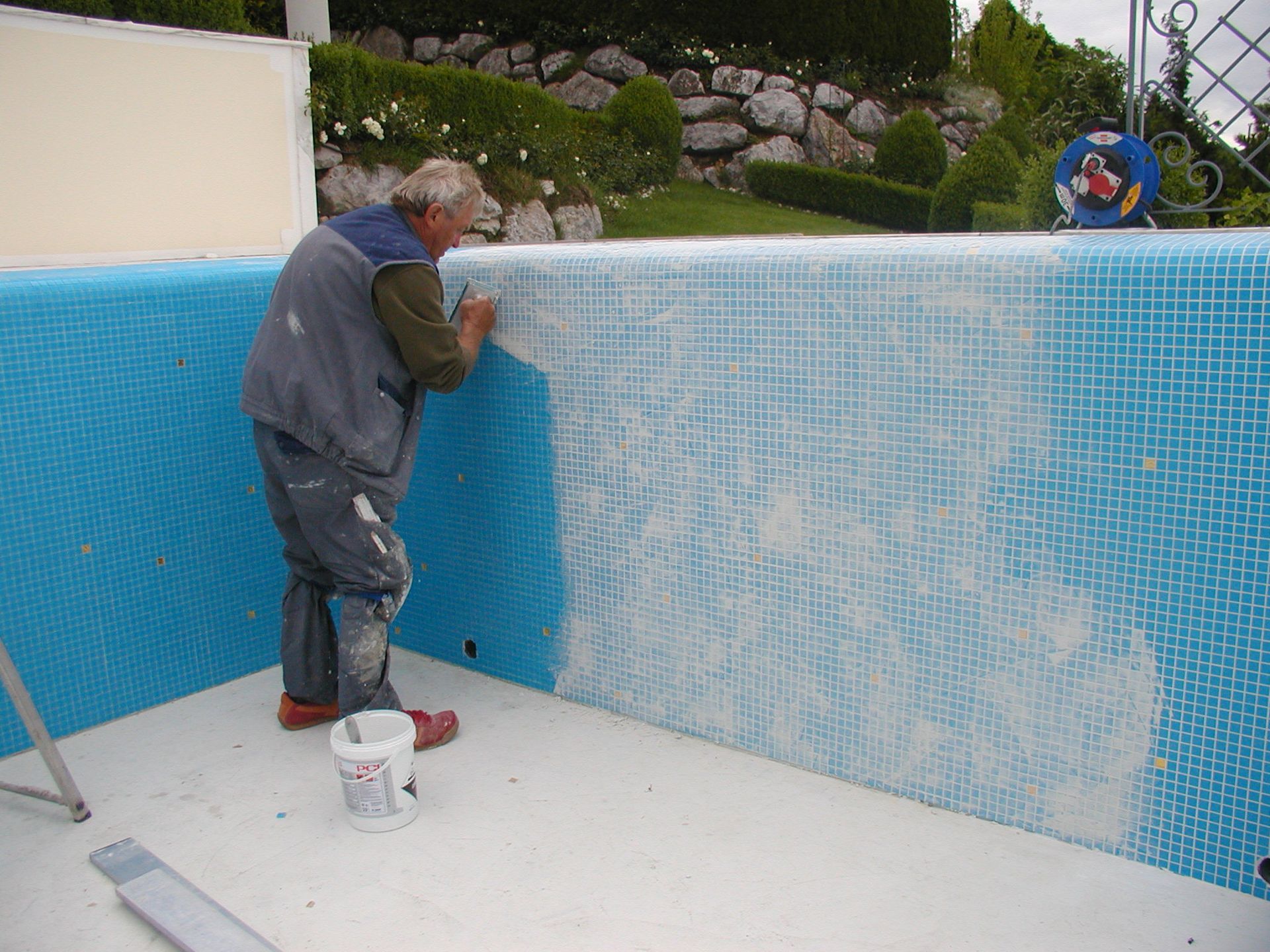 Ein Mann trägt Fugenmörtel auf blauen Fliesen in einem Pool auf. Er trägt farbbespritzte Kleidung und steht im Pool.