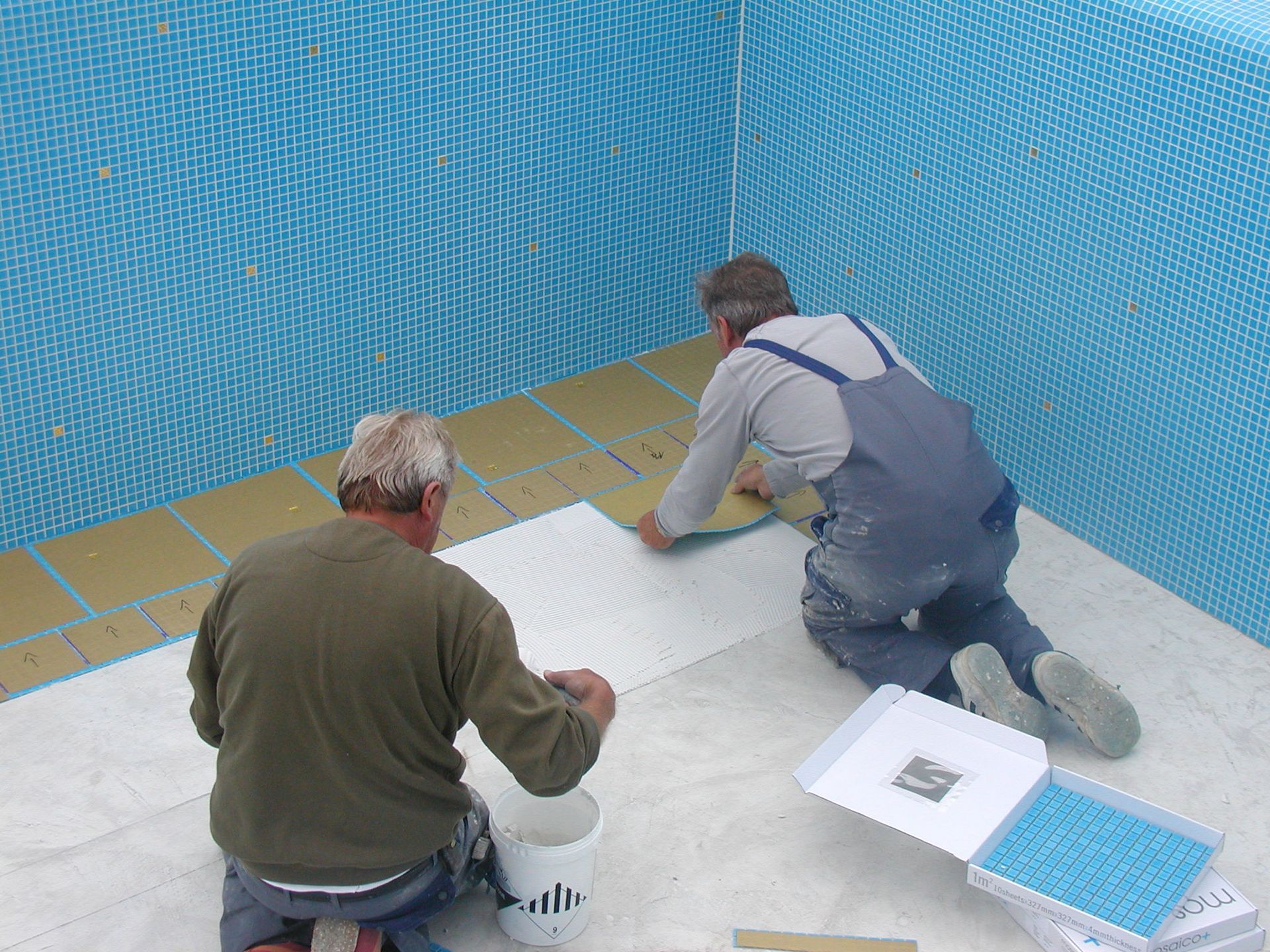 Zwei Männer installieren die Innenausstattung eines Pools mit blauen Fliesen; drinnen, kniend, bei der Arbeit.