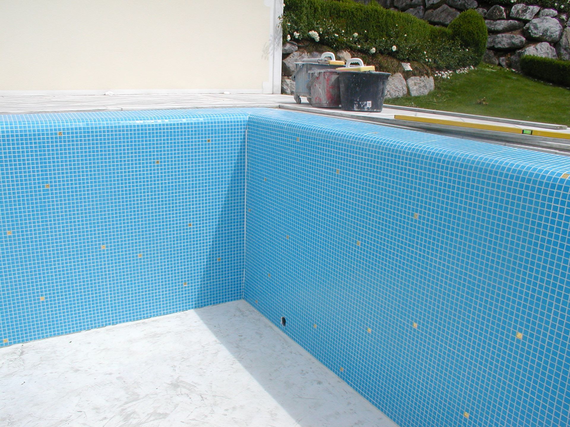 Im Bau befindlicher Swimmingpool mit blauen Fliesen, im Hintergrund Werkzeuge und Grün.