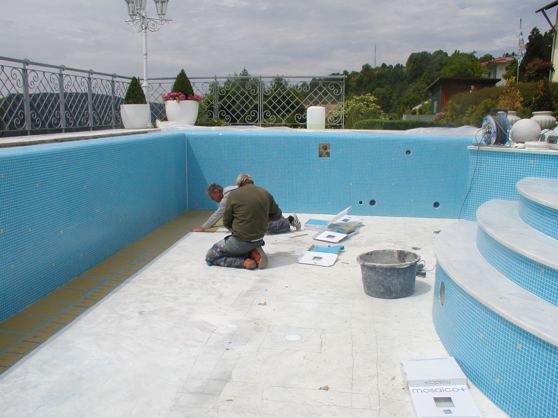 Zwei Arbeiter fliesen den Boden eines Schwimmbeckens. Türkis geflieste Wände, blauer Himmel, Außenanlage.