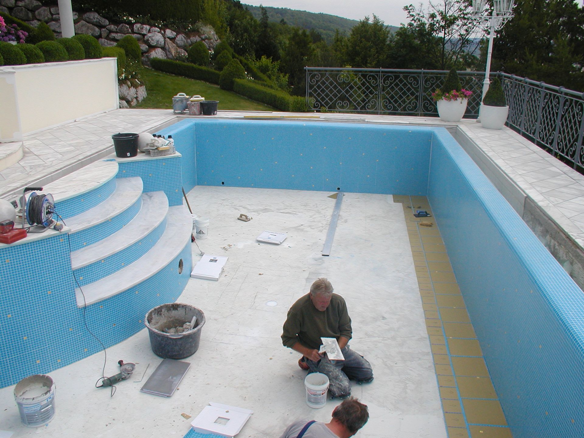 Männer fliesen einen im Bau befindlichen Pool mit blauen Fliesen; links Stufen, im Hintergrund Garten und Geländer.