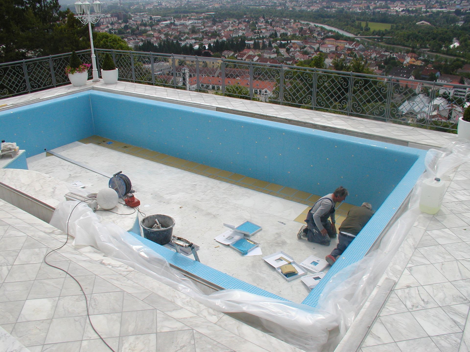 Zwei Arbeiter fliesen einen blau gefliesten Pool, im Hintergrund ist ein Blick auf die Stadt zu sehen.