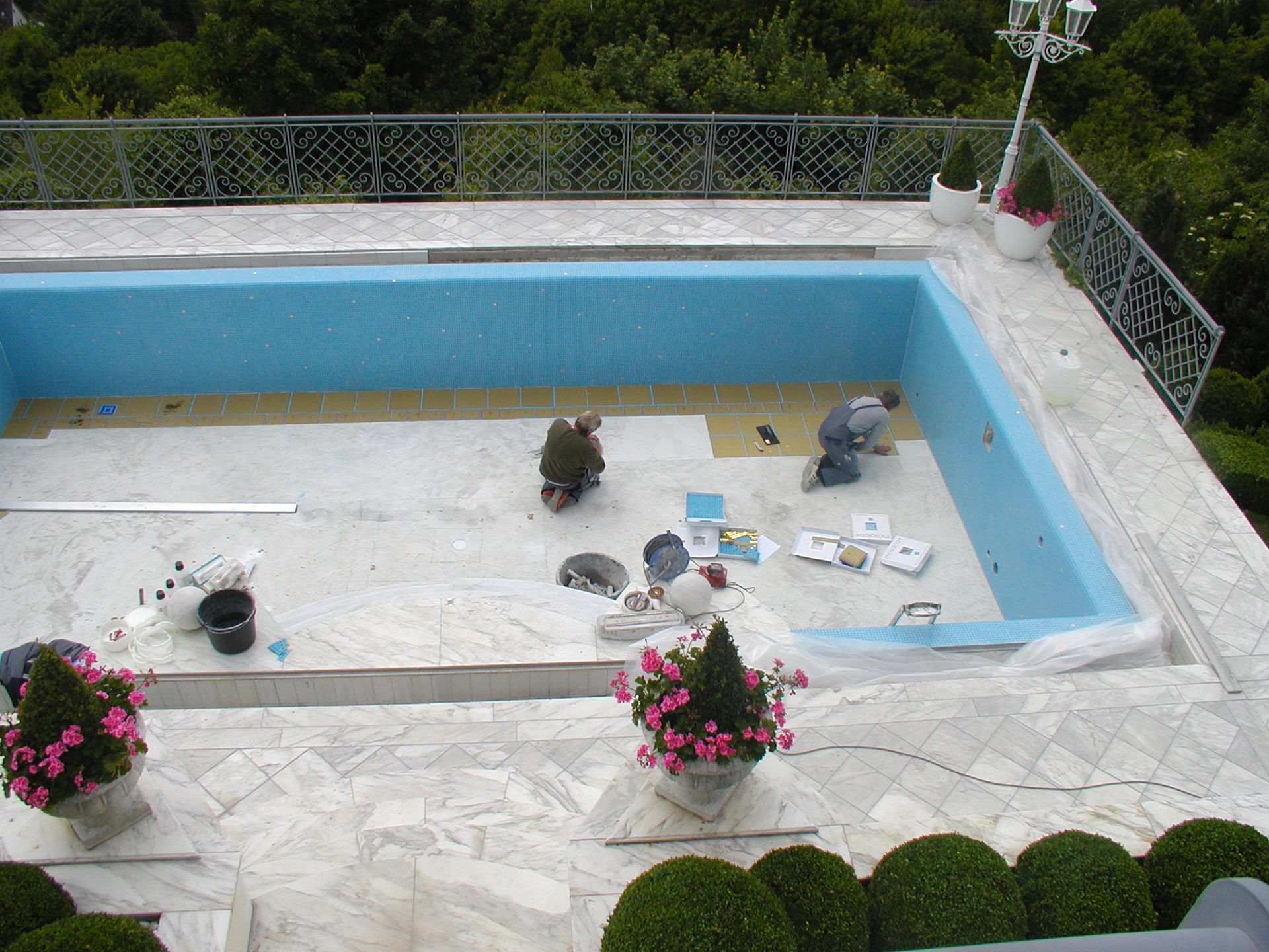 Arbeiter fliesen einen blauen Pool, der auf einer weißen Steinterrasse liegt und von Grün umgeben ist.