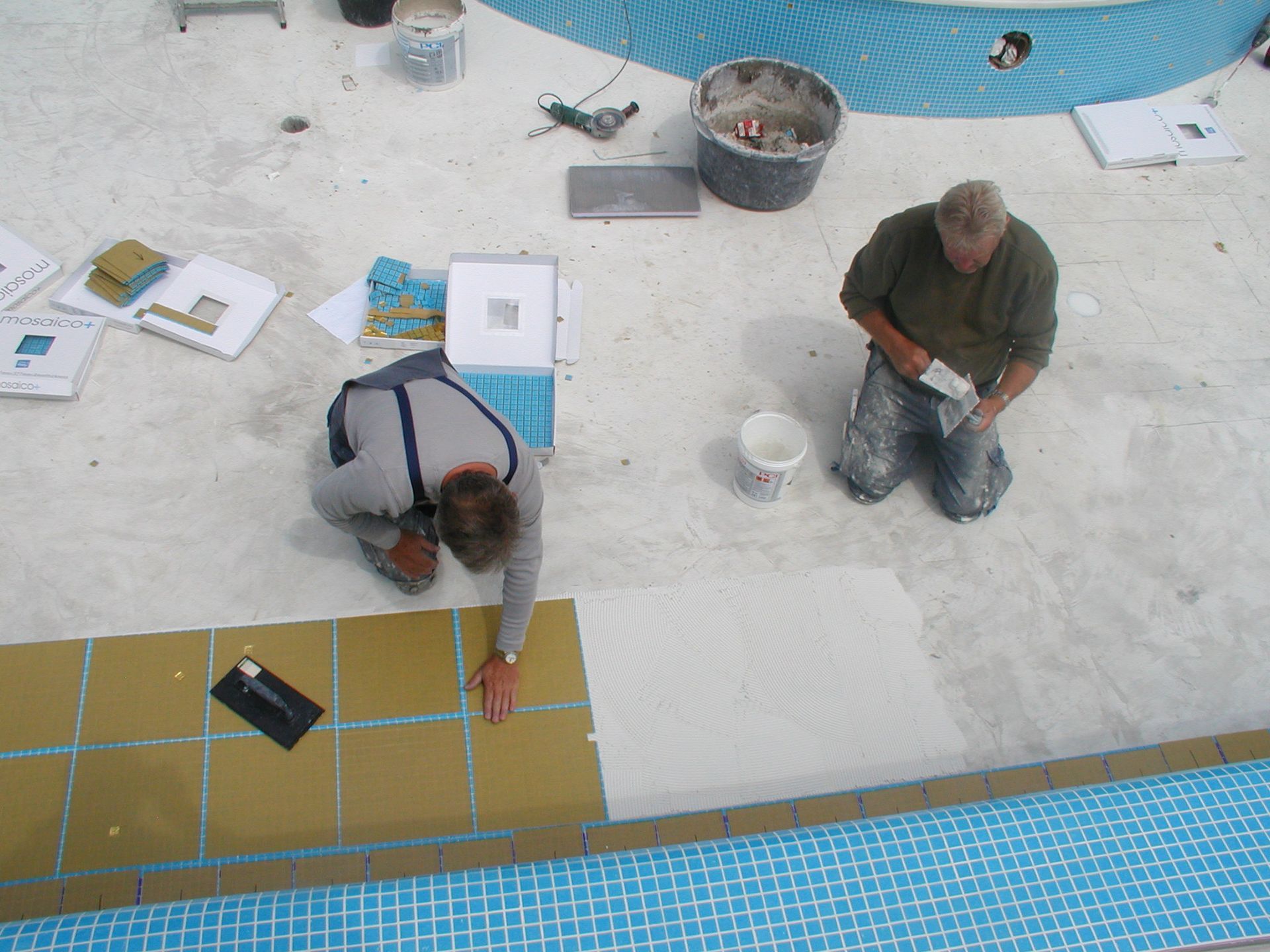 Zwei Männer fliesen einen Pool: Einer verteilt Fugenmörtel, der andere legt Fliesen auf den Poolboden.
