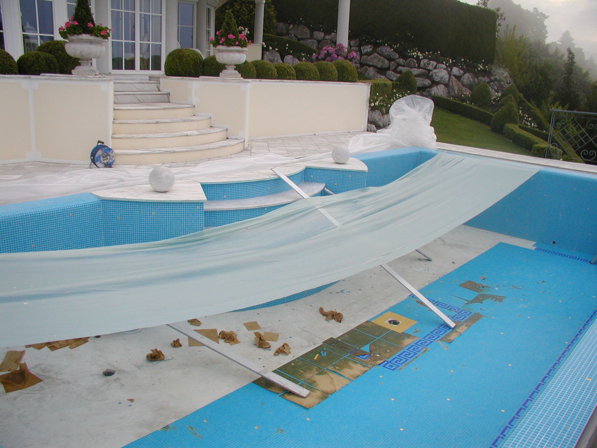 Pool mit zurückgezogener Abdeckung, die blaue Fliesen und Schutt freigibt, vor einem Haus.