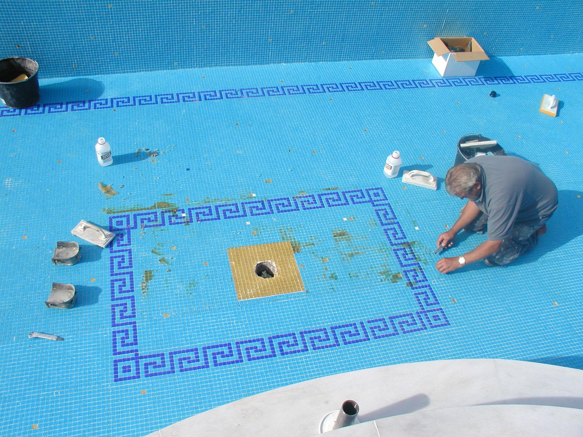 Eine Person repariert in einer sonnigen Umgebung ein Poolmosaik mit blauen und goldenen Fliesen.