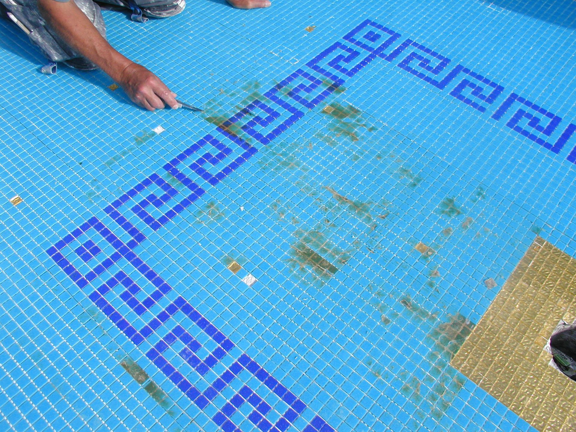 Person repariert einen blau gefliesten Pool mit griechischem Schlüsselranddesign.