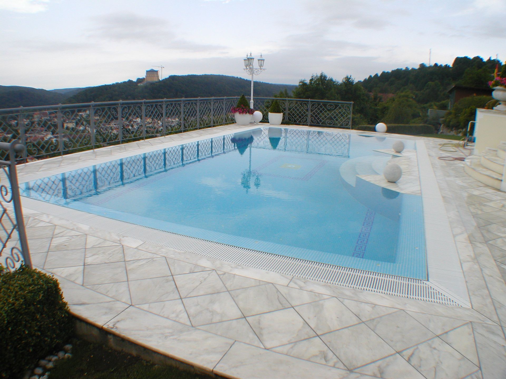 Ein rechteckiger Swimmingpool mit blauem Wasser und weißem Marmorboden mit Blick auf eine Stadt und Hügel.
