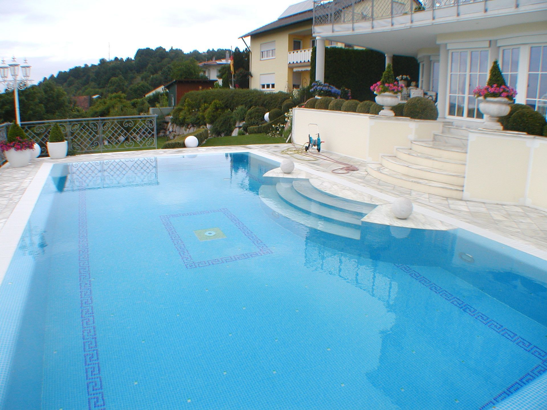 Swimmingpool mit blauem Wasser und Mosaikfliesenverkleidung, neben einem Haus mit Balkon, Stufen und grüner Landschaftsgestaltung.