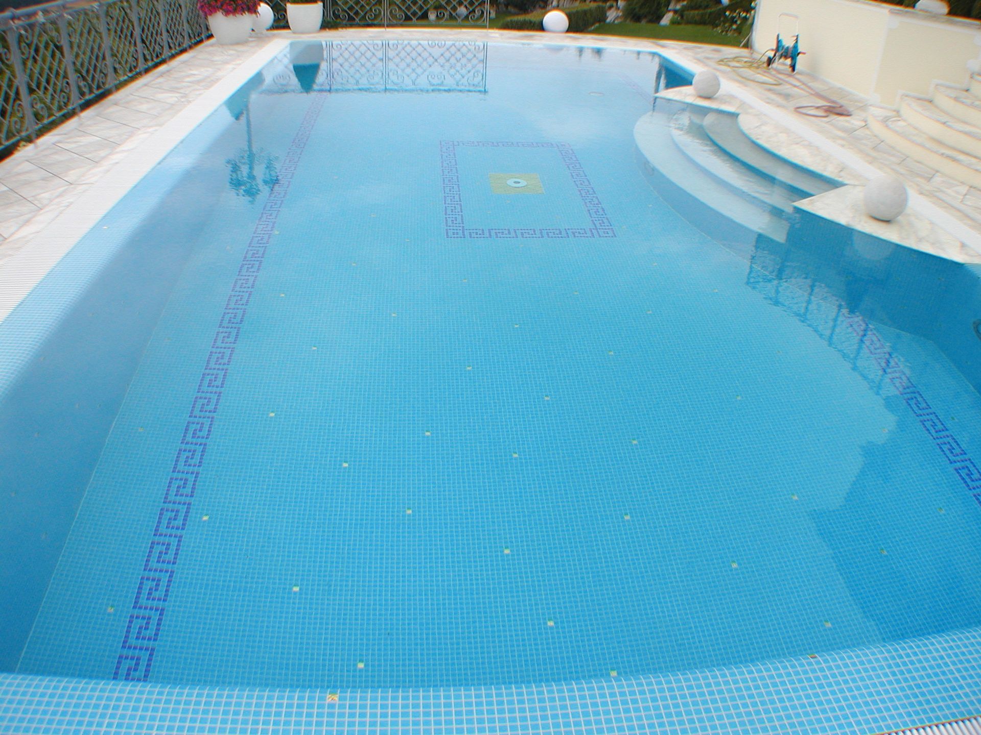 Blau gefliester Swimmingpool mit Stufen und dekorativer Umrandung, umgeben von weißen Steinen und Geländer.