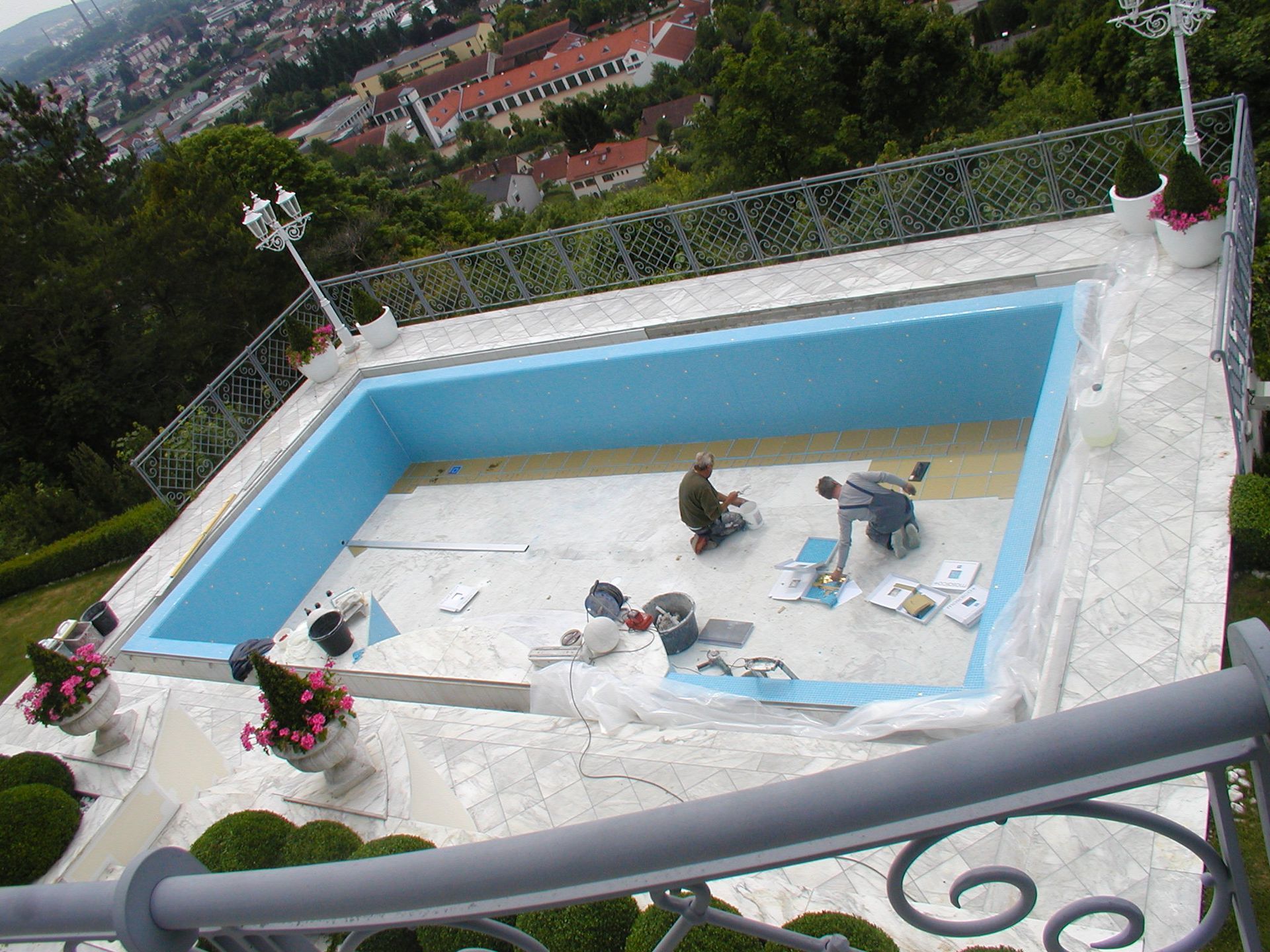 Arbeiter tragen Dichtungsmittel auf ein leeres, hellblaues Schwimmbecken auf und blicken auf die Stadtlandschaft.