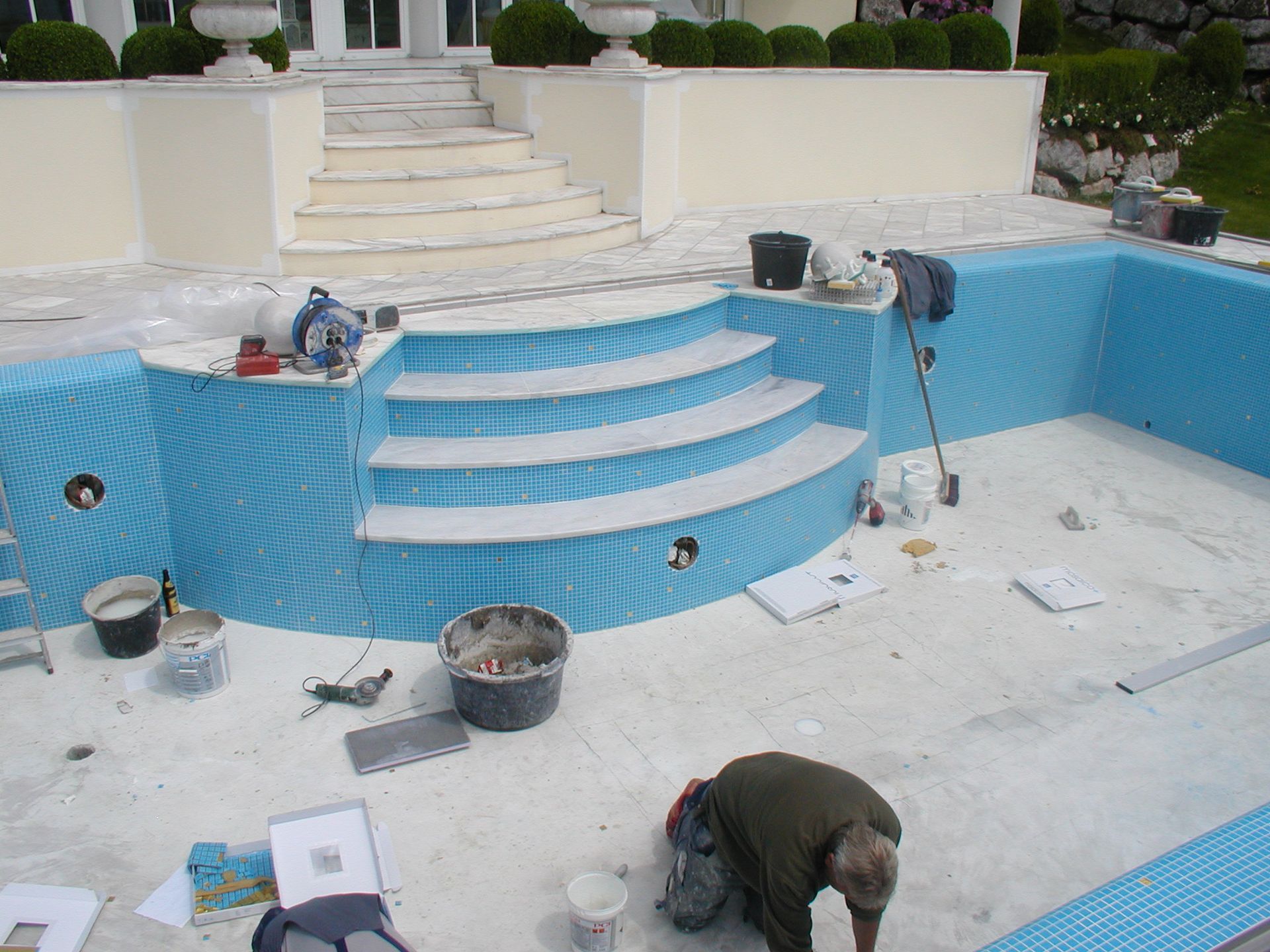 Pool im Bau; blau geflieste Wände, weiße Stufen, Arbeiter beim Fliesenlegen.