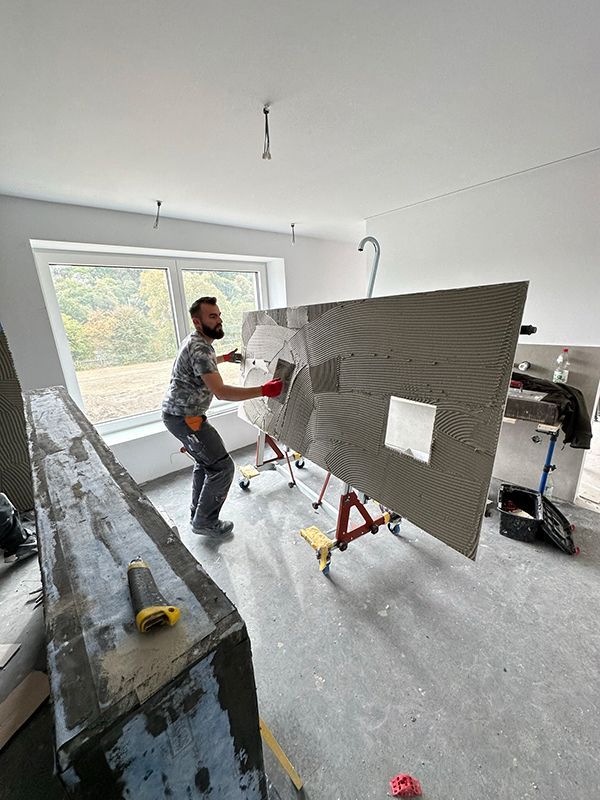 Mann trägt mit einem Rollwagen in einem Raum mit großen Fenstern Klebstoff auf eine große Fliese auf.