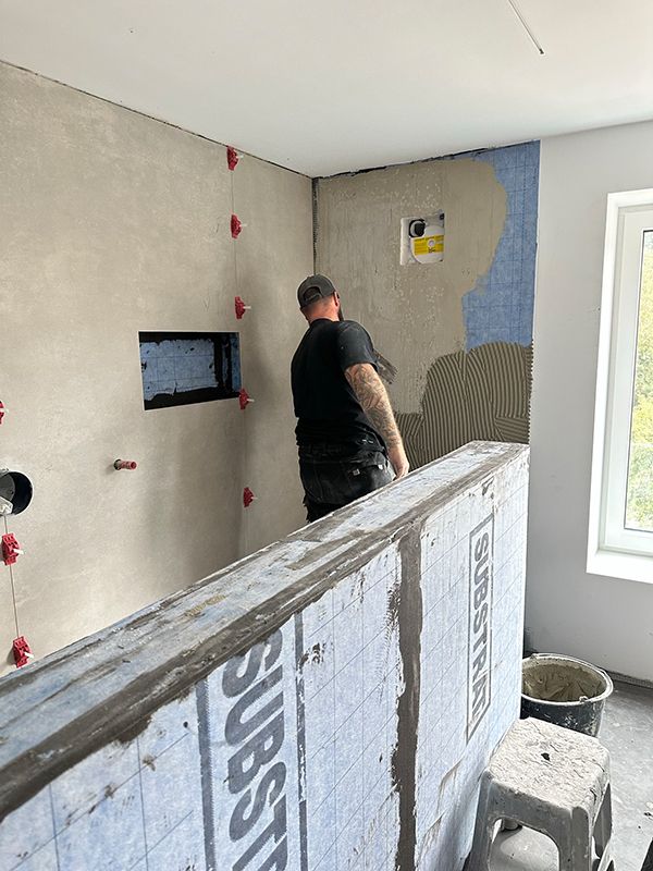 Ein Mann fliest ein Badezimmer. Graue Wände, blaue Feuchtigkeitssperre, rote Fliesenabstandshalter.