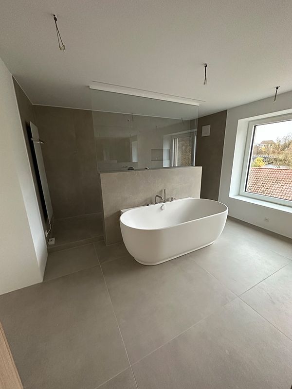 Modernes Badezimmer mit weißer Badewanne, Dusche hinter Glas, Fenster und grauem Fliesenboden.