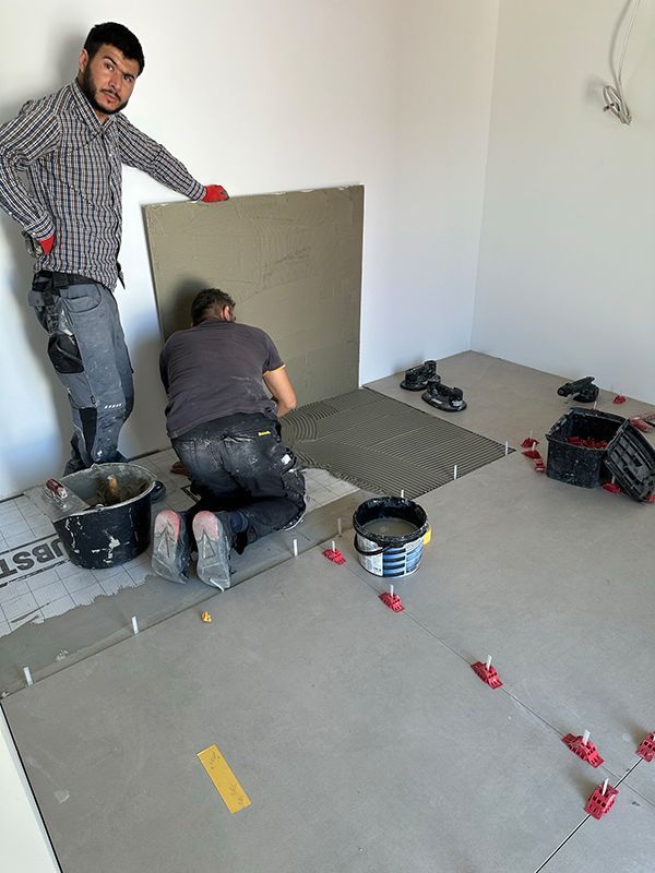 Zwei Männer fliesen einen Raum; einer hält eine große Fliese, der andere kniet und trägt Klebstoff auf. Werkzeuge und Materialien liegen verstreut herum.