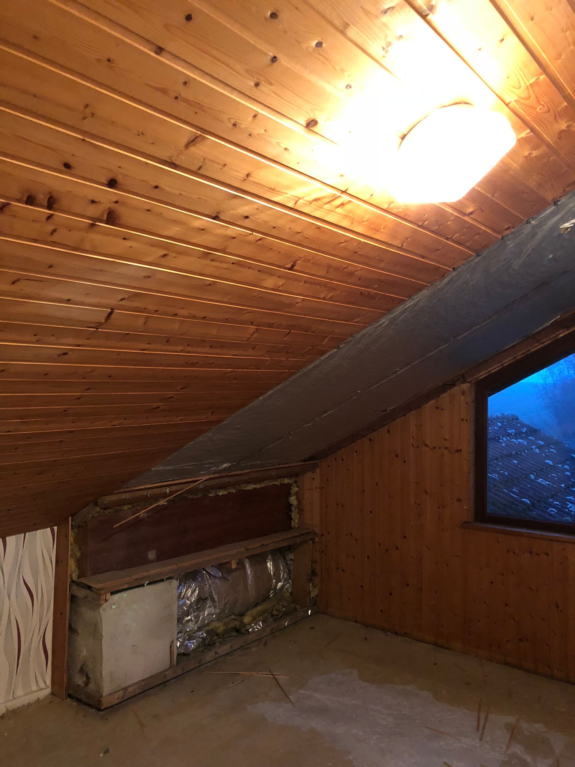 Dachzimmer mit Wänden und Decke aus Holz, einem kleinen Fenster und einem quer verlaufenden silbernen Kanal.
