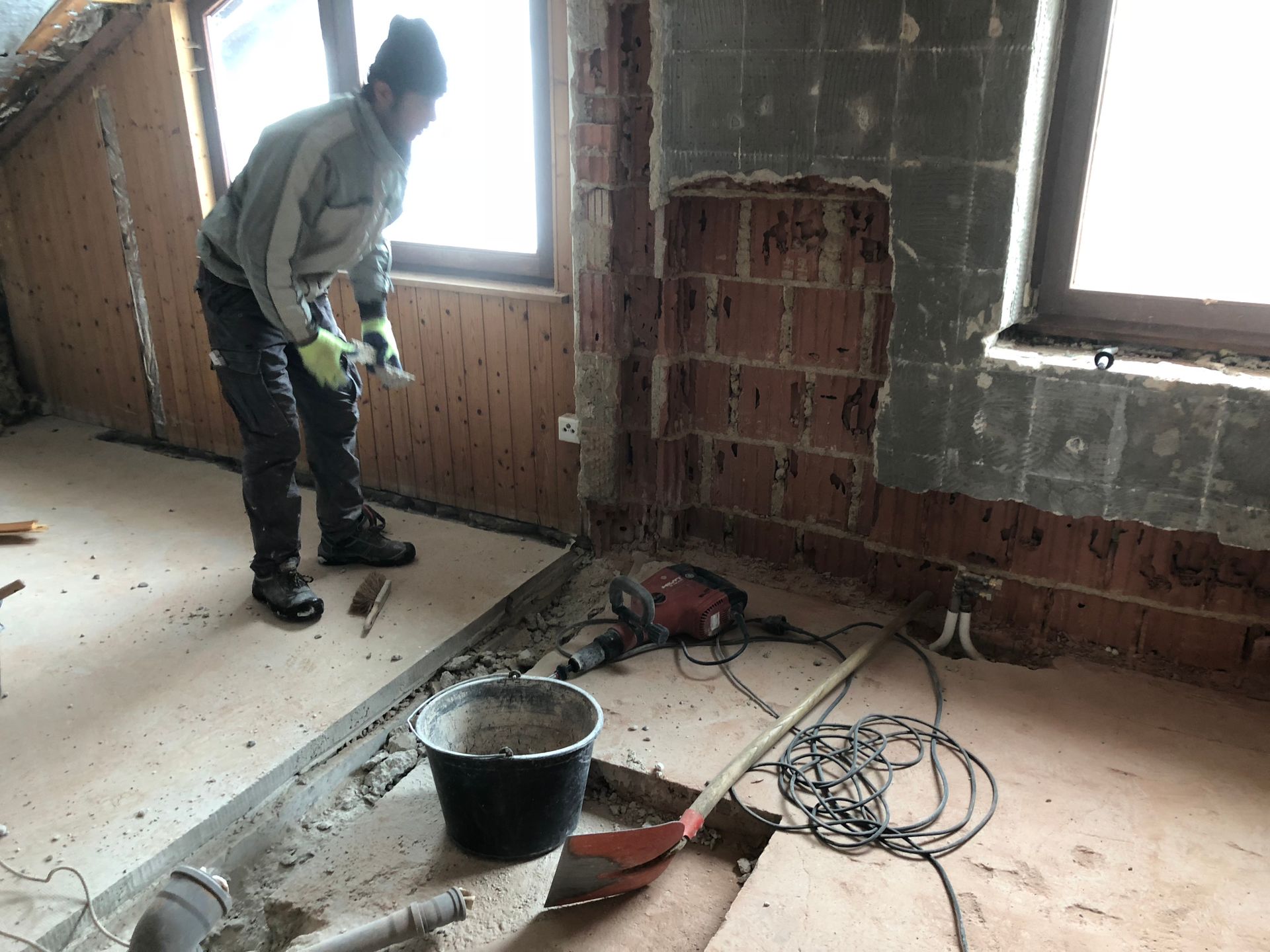 Eine Person, die während Bauarbeiten mit einem Elektrowerkzeug an einer Ziegelwand in einem Raum mit Fenster arbeitet.