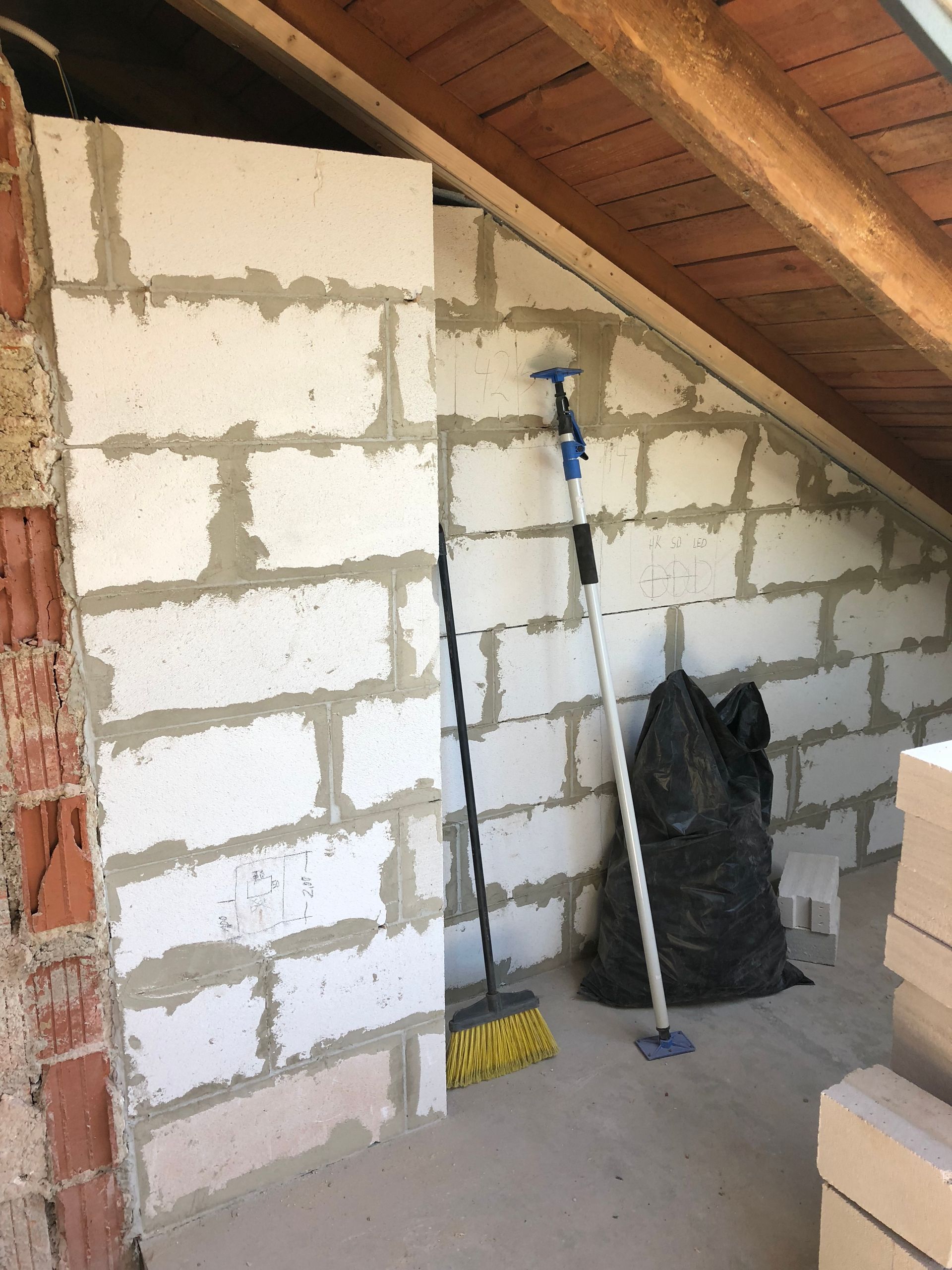 Unfertige Innenwand mit Schlackenblöcken, einem Besen und einem Müllsack unter einem schrägen Dach.
