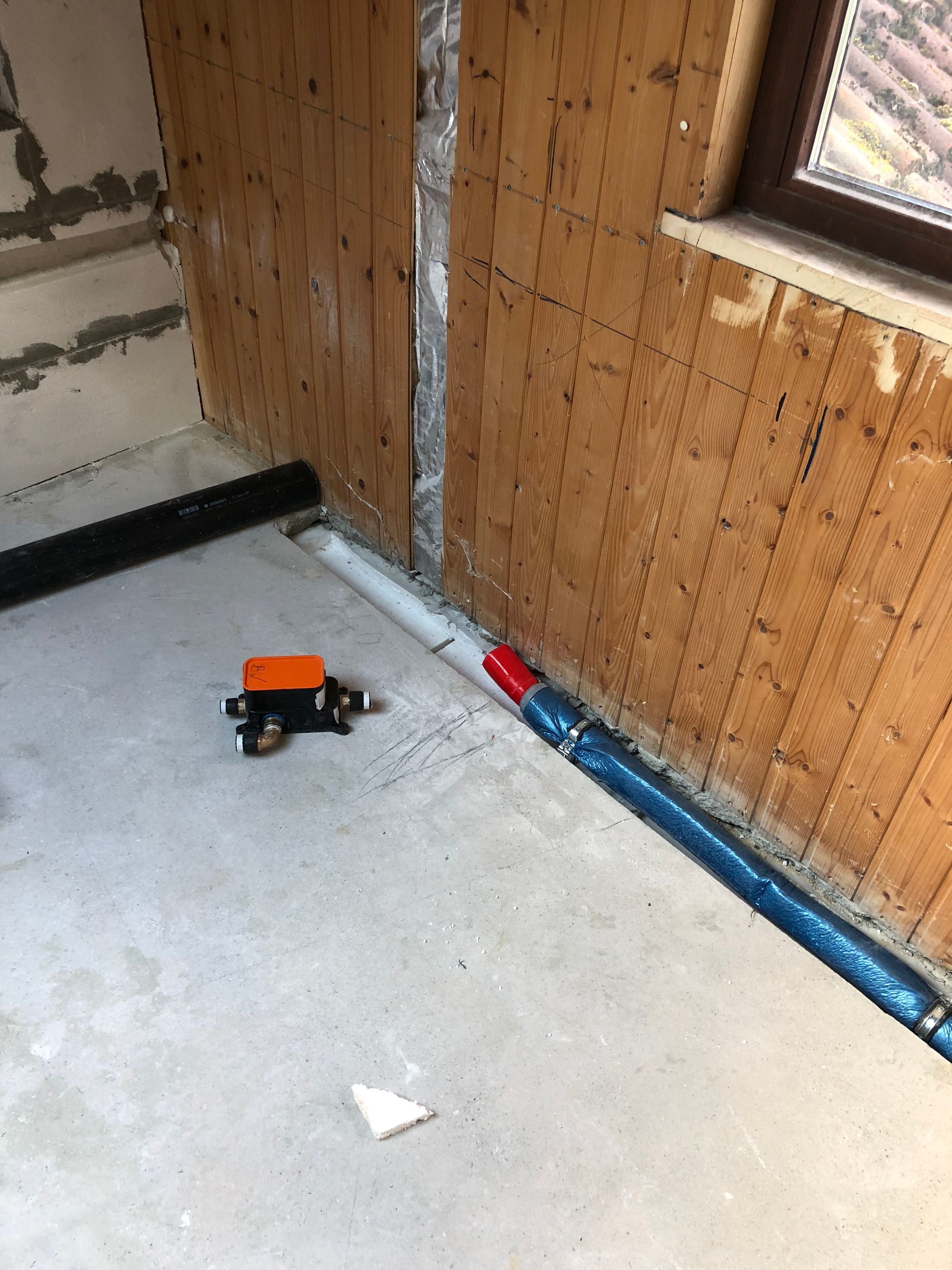 Betonboden, freiliegende Holzwände, Rohre und ein kleines schwarz-oranges Objekt, möglicherweise ein Ventil, in einem im Bau befindlichen Raum.