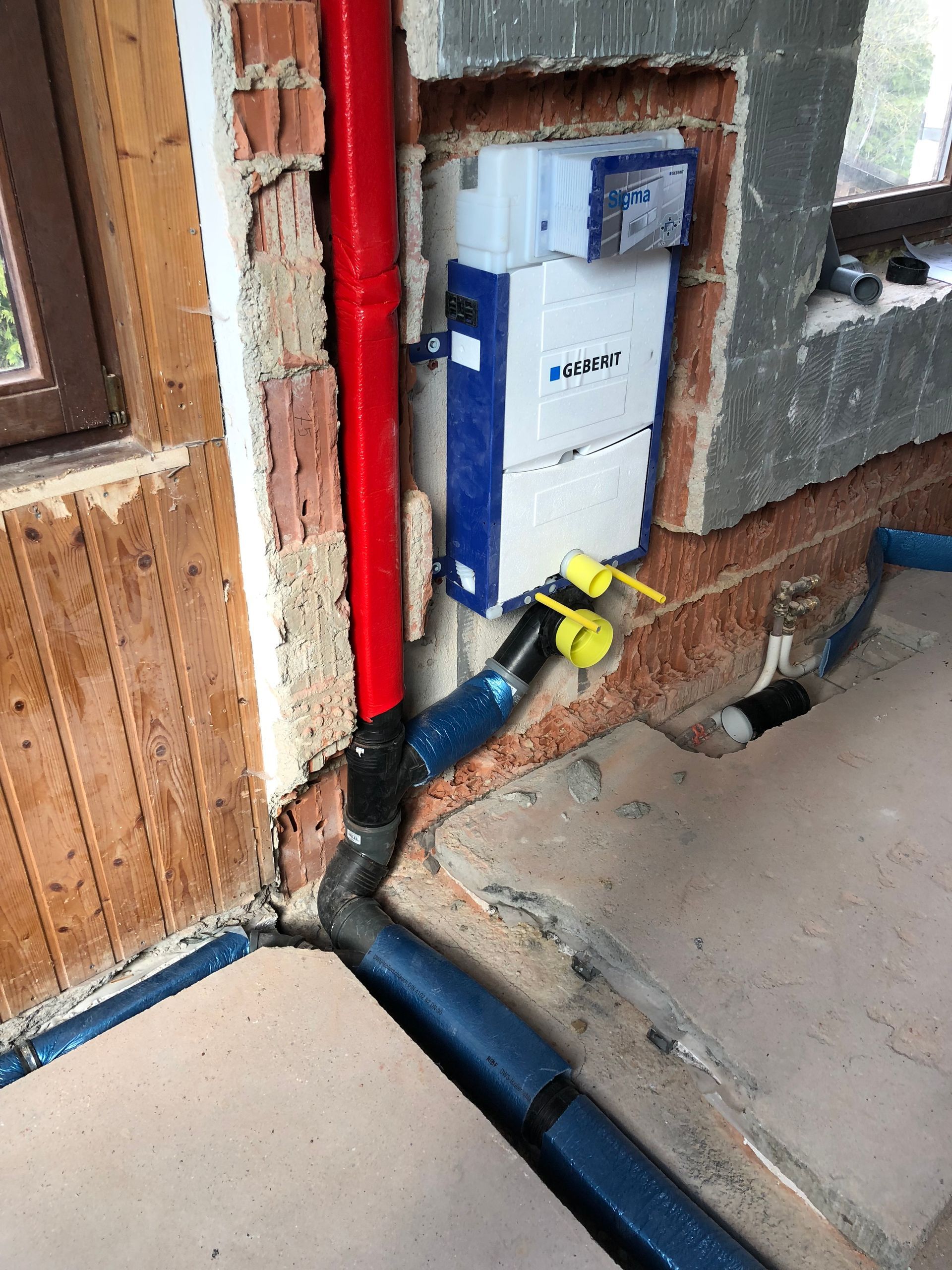Sanitärinstallation in einem Wandhohlraum: rotes Rohr, blaue isolierte Rohre, weiße Spüleinheit, gelbe Absperrventile, Ziegelwand.