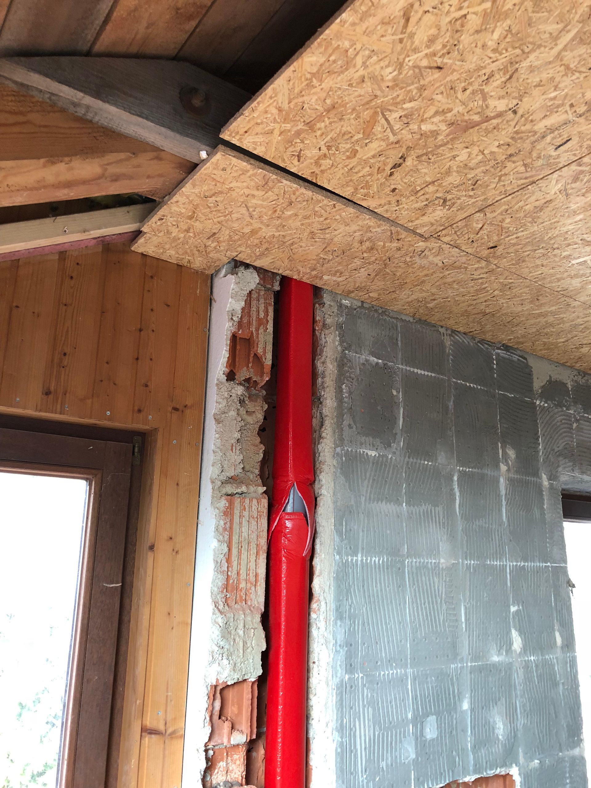 Ein rotes Rohr verläuft vertikal zwischen Sichtziegeln und einer grau gefliesten Wand, in der Nähe einer Holztür und einer teilweise konstruierten Decke.