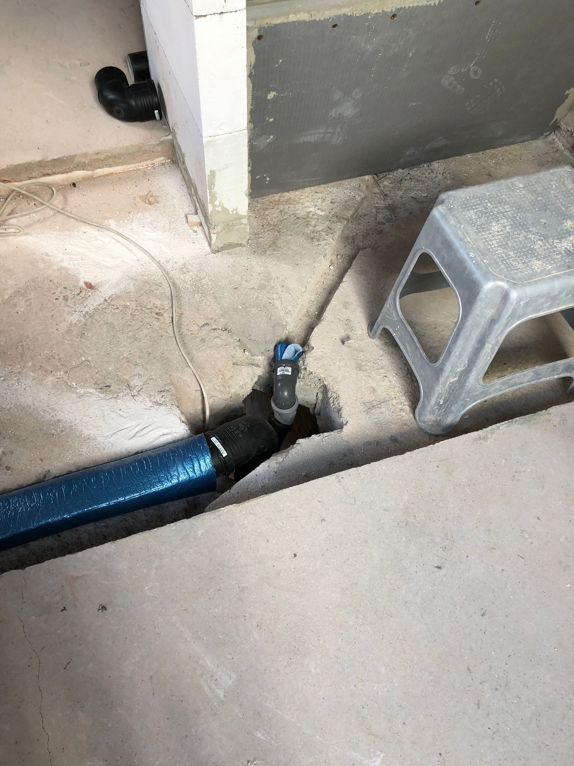 Ein blauer Schlauch und ein schwarzes Rohr ragen aus einer Betonöffnung in der Nähe eines kleinen grauen Hockers.