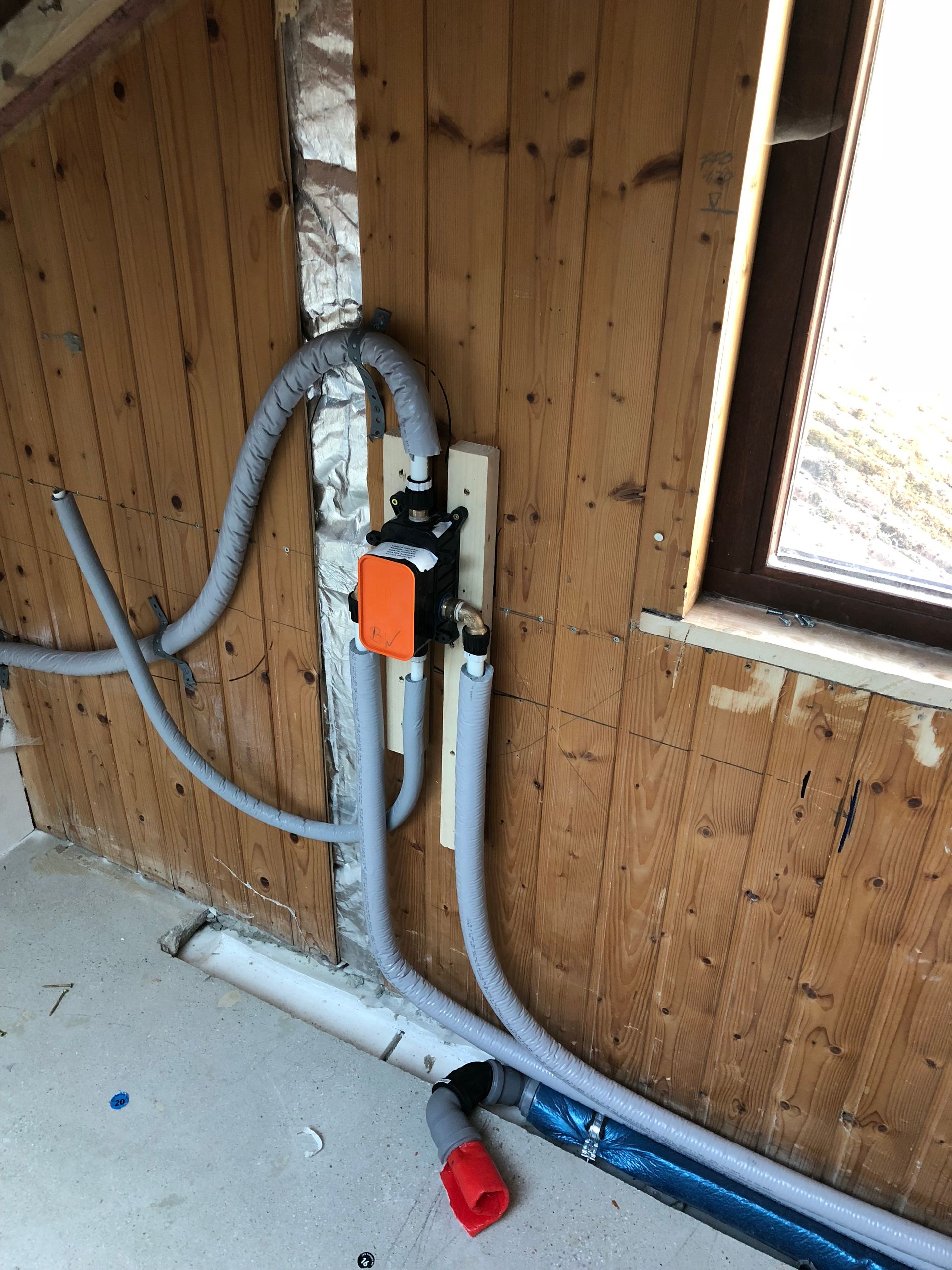 Sanitärinstallation an einer Holzwand. Schwarze Pumpe, graue Schläuche, weiße Rohre und eine rote Kappe.