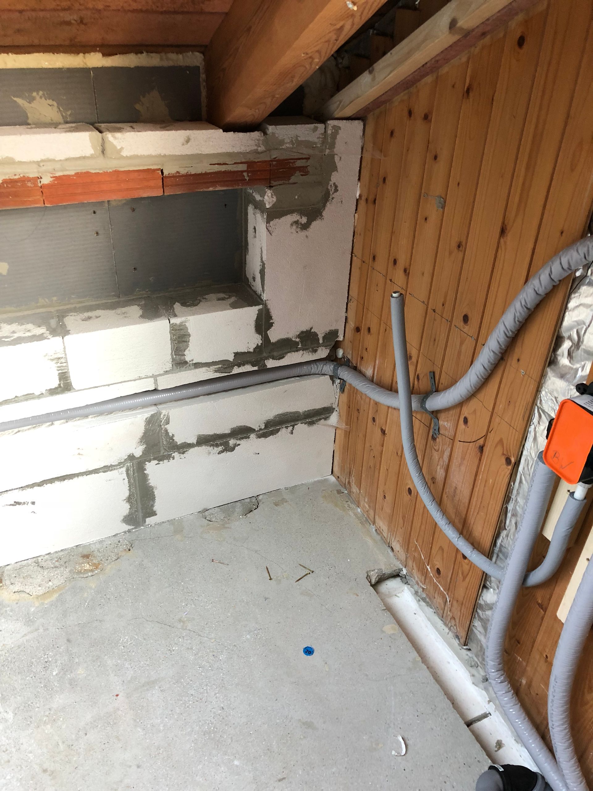 Graue Leitungsrohre an einer Betonblockwand, einer Holzwand und einem Betonboden. Orangefarbener Schaltkasten.