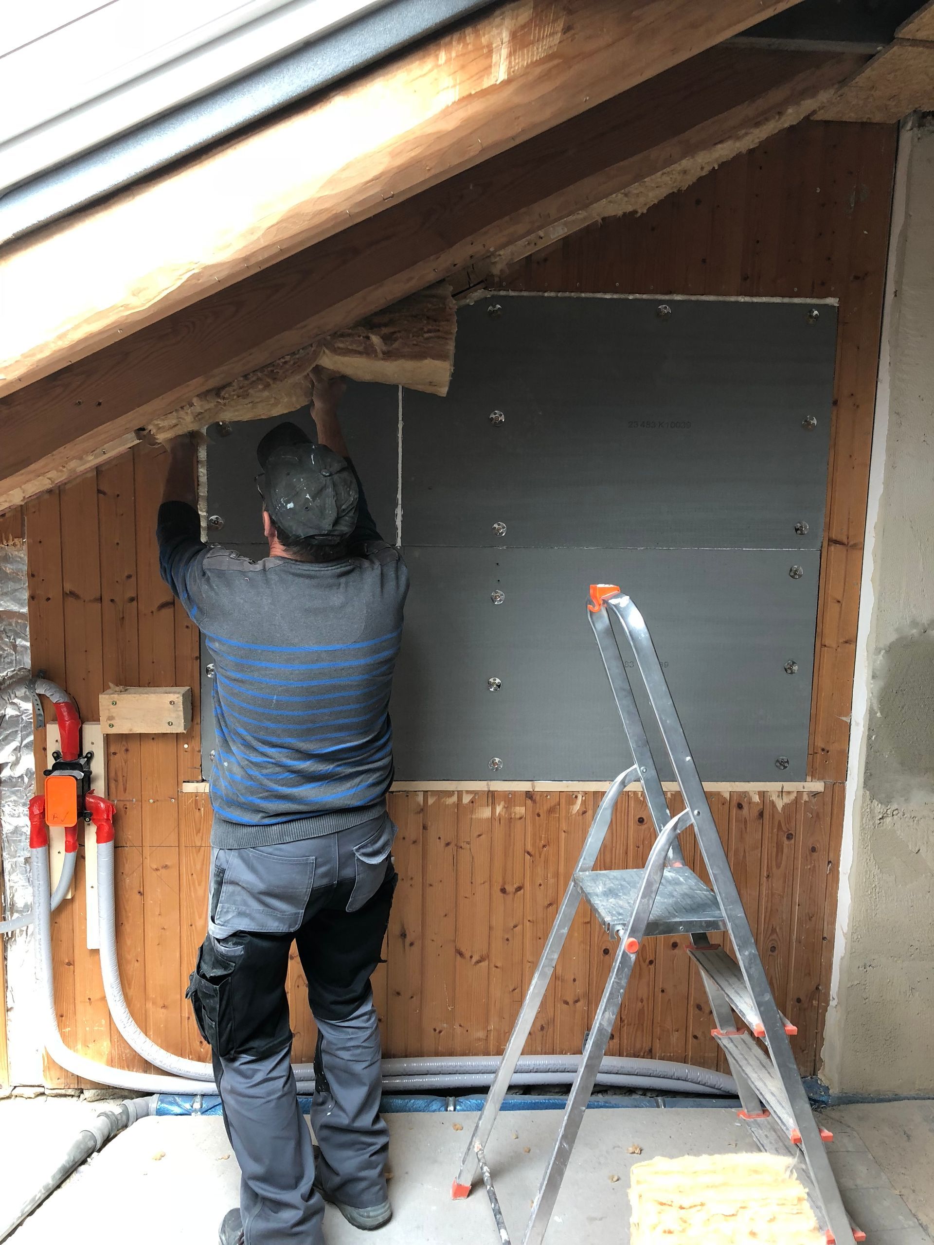 Eine Person installiert eine dunkle Platte an einer Holzwand, neben einer Leiter, Rohren und einem Fenster.