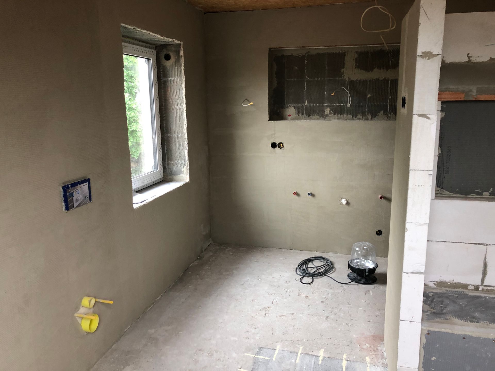 Innenansicht einer Baustelle: Ein Raum mit verputzten Wänden, Fenster und freiliegenden elektrischen Leitungen.