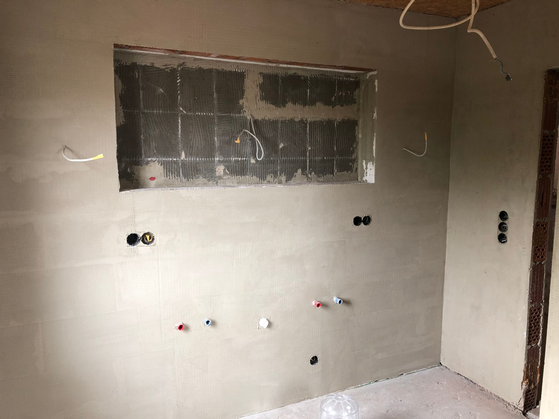 Innenwand in einem im Bau befindlichen Raum; grauer Putz, eine Fensteröffnung und Elektro-/Sanitärinstallationen.