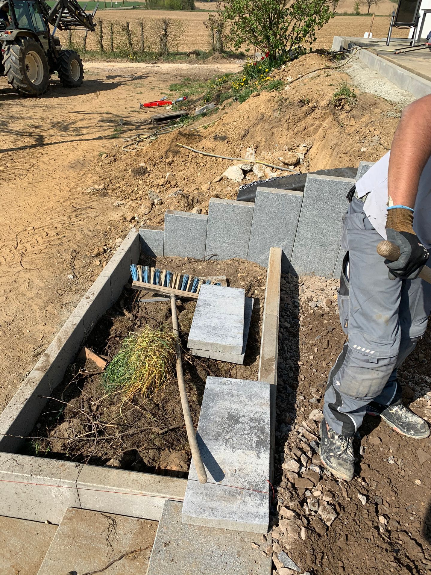 Bau von Steinstufen, ein Hochbeet. Eine Person hält einen Rechen und ein Traktor ist zu sehen.