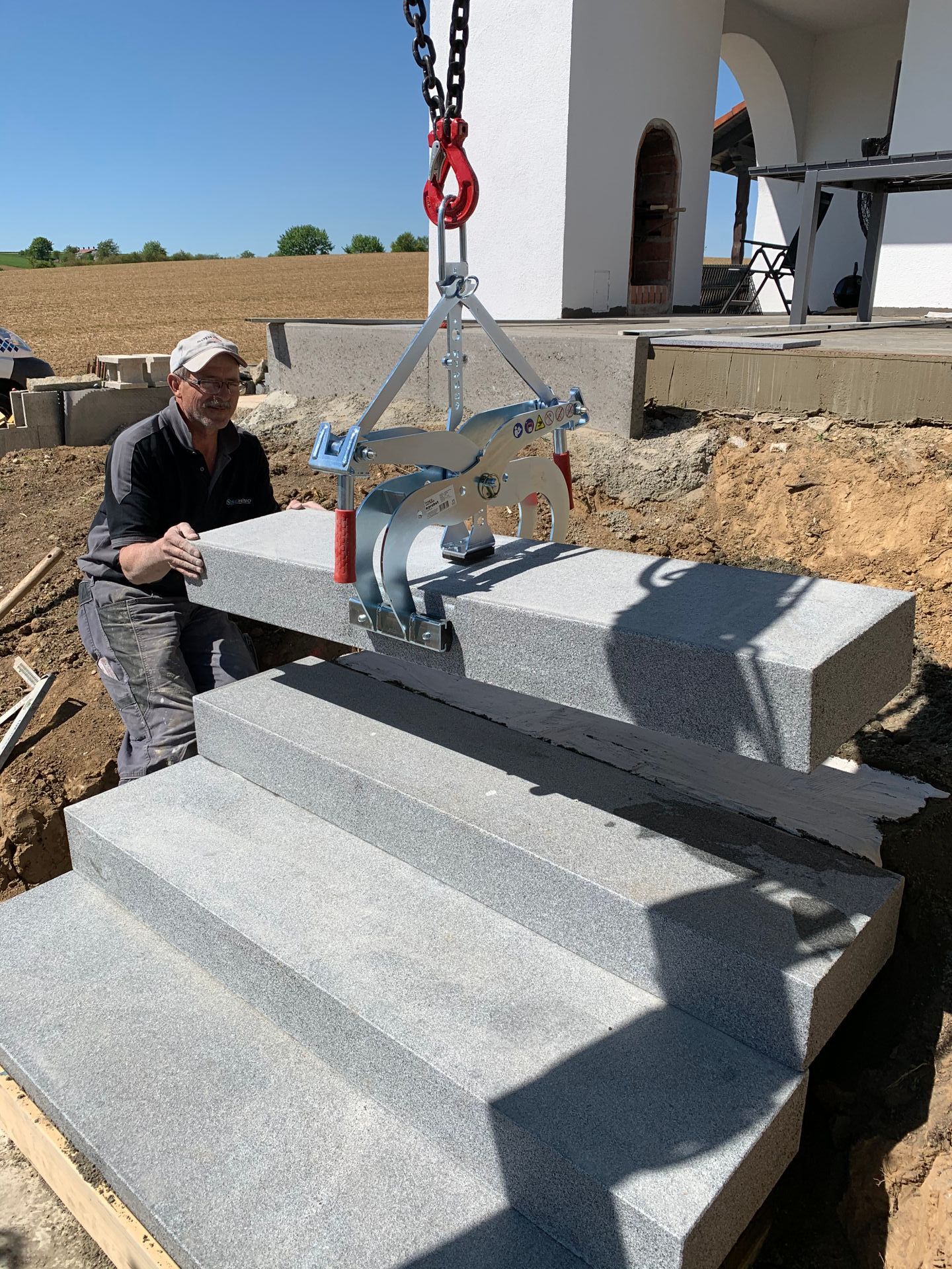 Bauarbeiter verwendet einen Hebezeug, um eine Granitstufe zu platzieren. Draußen, heller Tag.