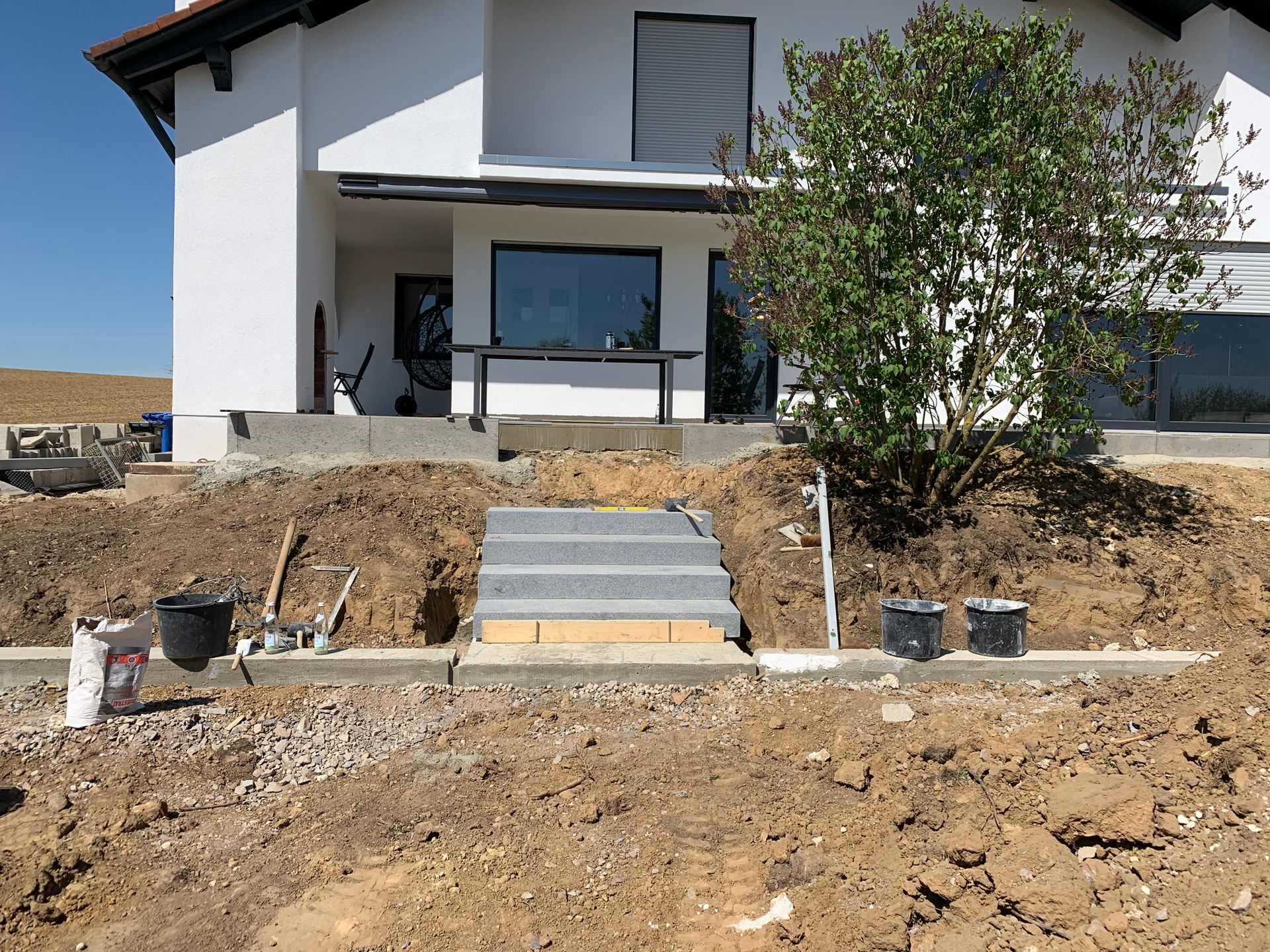 Außenansicht eines im Bau befindlichen Hauses, Erdhügel, Steinstufen, unfertige Landschaftsgestaltung und ein weißes Gebäude.