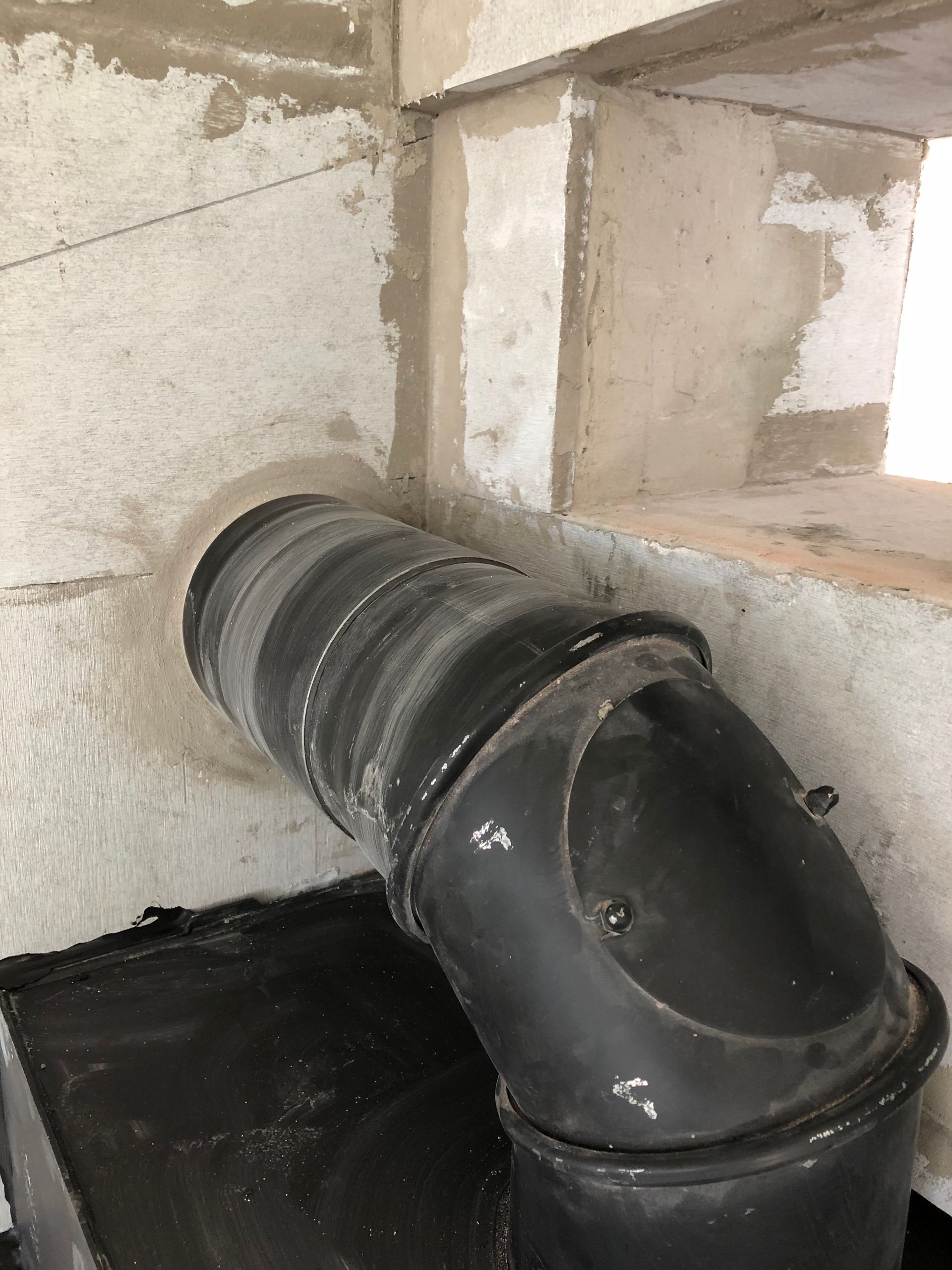 Schwarzer Rohrbogen, der aus einer leichten Betonwand herausragt, wahrscheinlich ein Schornstein für einen Holzofen.