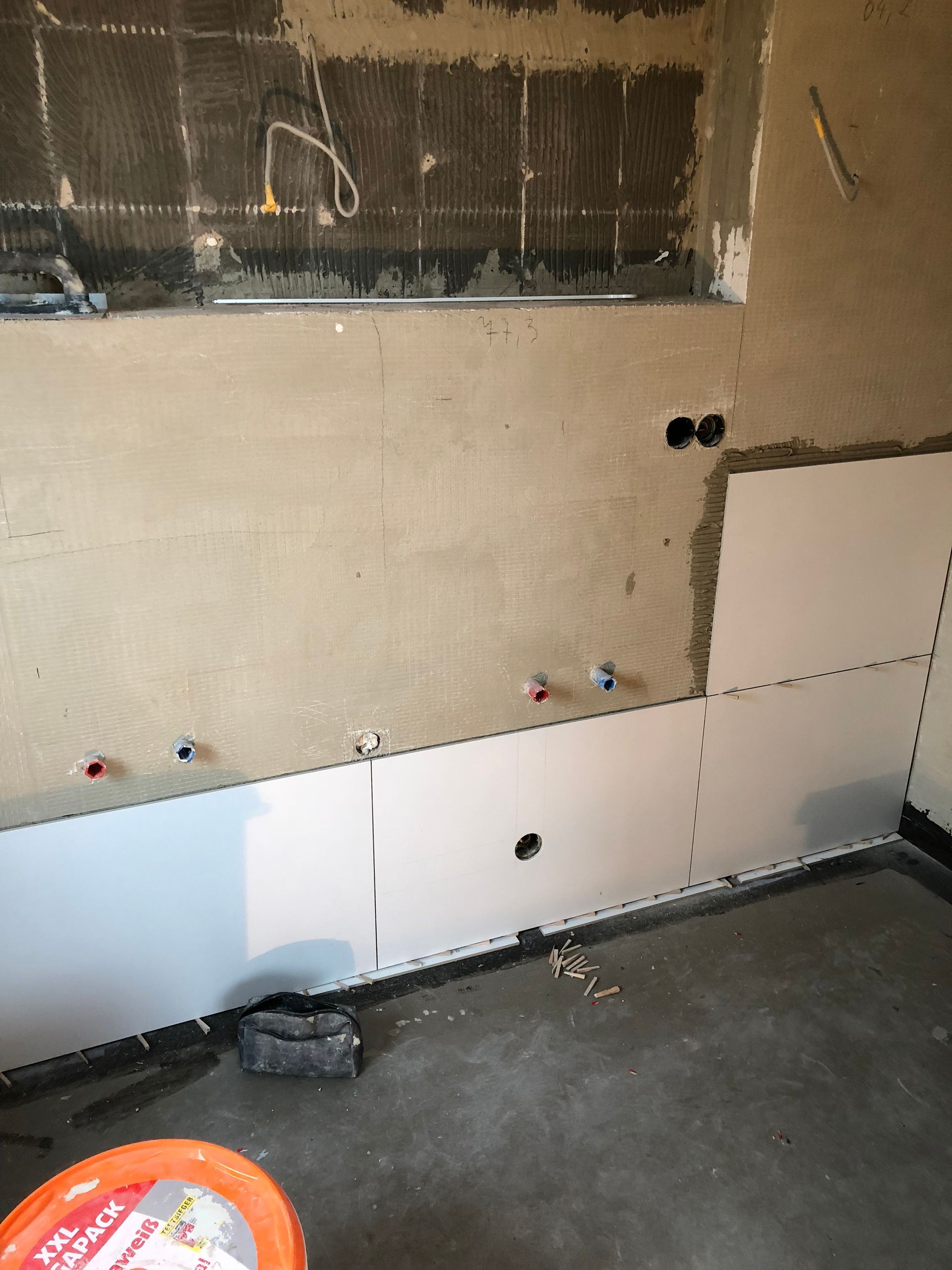 Baustelle mit teilweise gefliesten Wänden. Rohre und Leitungen liegen frei. Orangefarbener Eimer und Werkzeuge auf dem Boden.