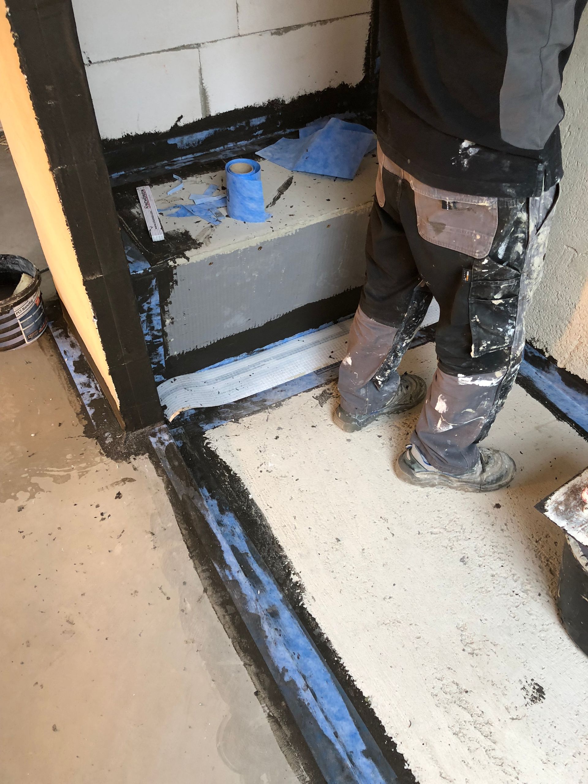 Person, die in einem Betonraum schwarzes Dichtungsmittel um einen Duschabfluss und eine Türöffnung aufträgt.