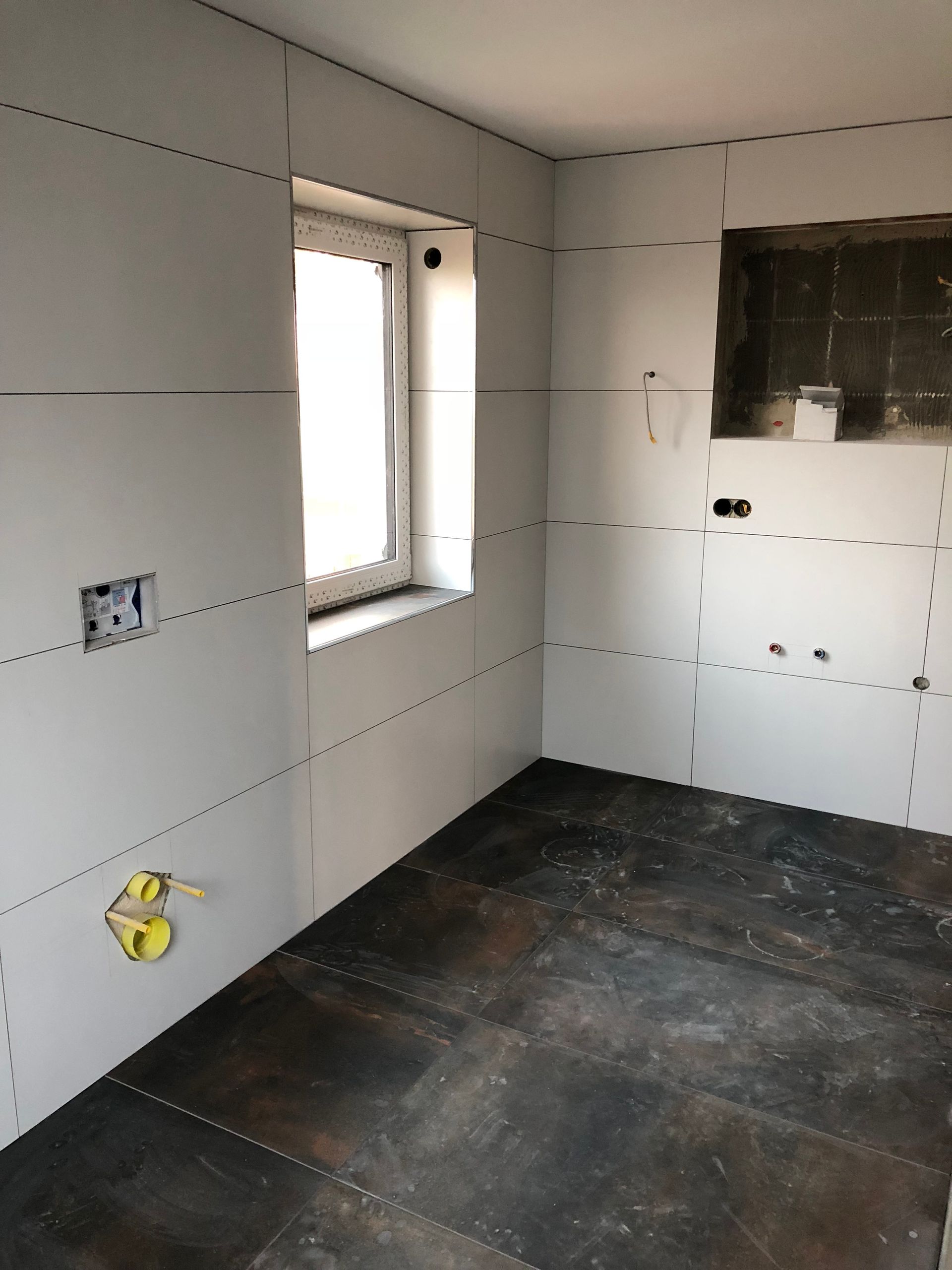 Innenansicht eines im Bau befindlichen Raumes mit Trockenbauwänden und einem Fenster, der Boden ist mit dunklem Material bedeckt.