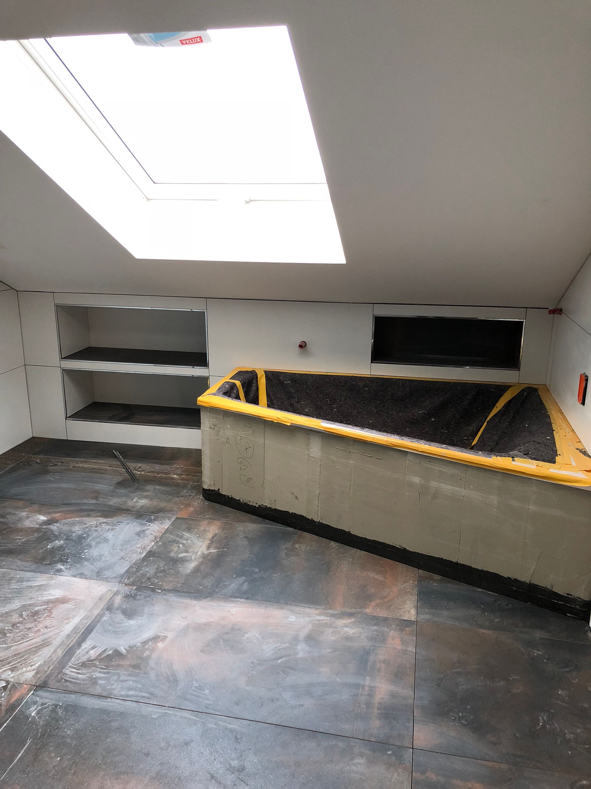Unfertiger Raum mit Oberlicht, eingebauten Regalen und einer großen rechteckigen Betonkonstruktion mit gelben Verzierungen.