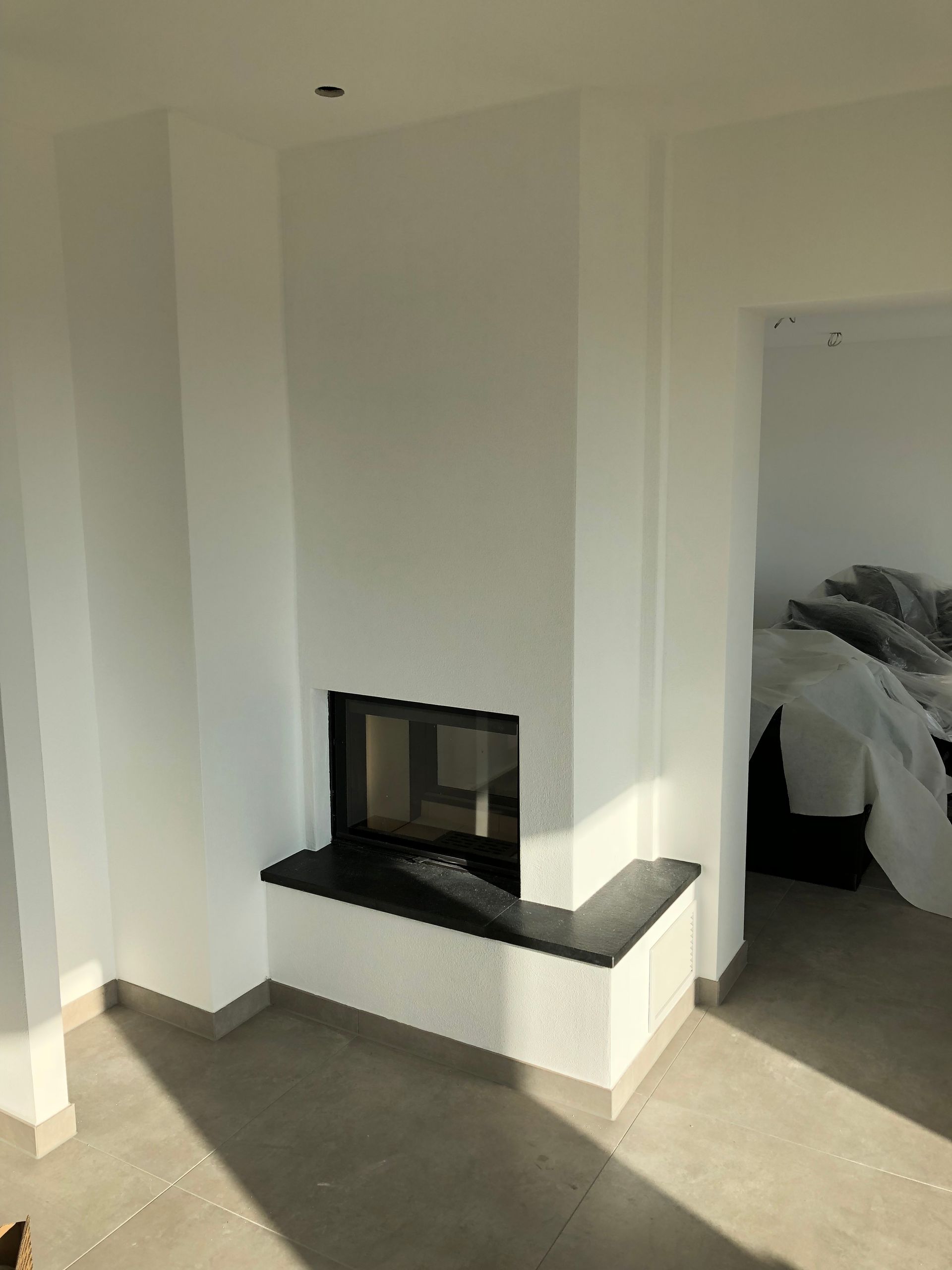 Weißer Kamin mit schwarzer Feuerstelle in einem lichtdurchfluteten Raum.