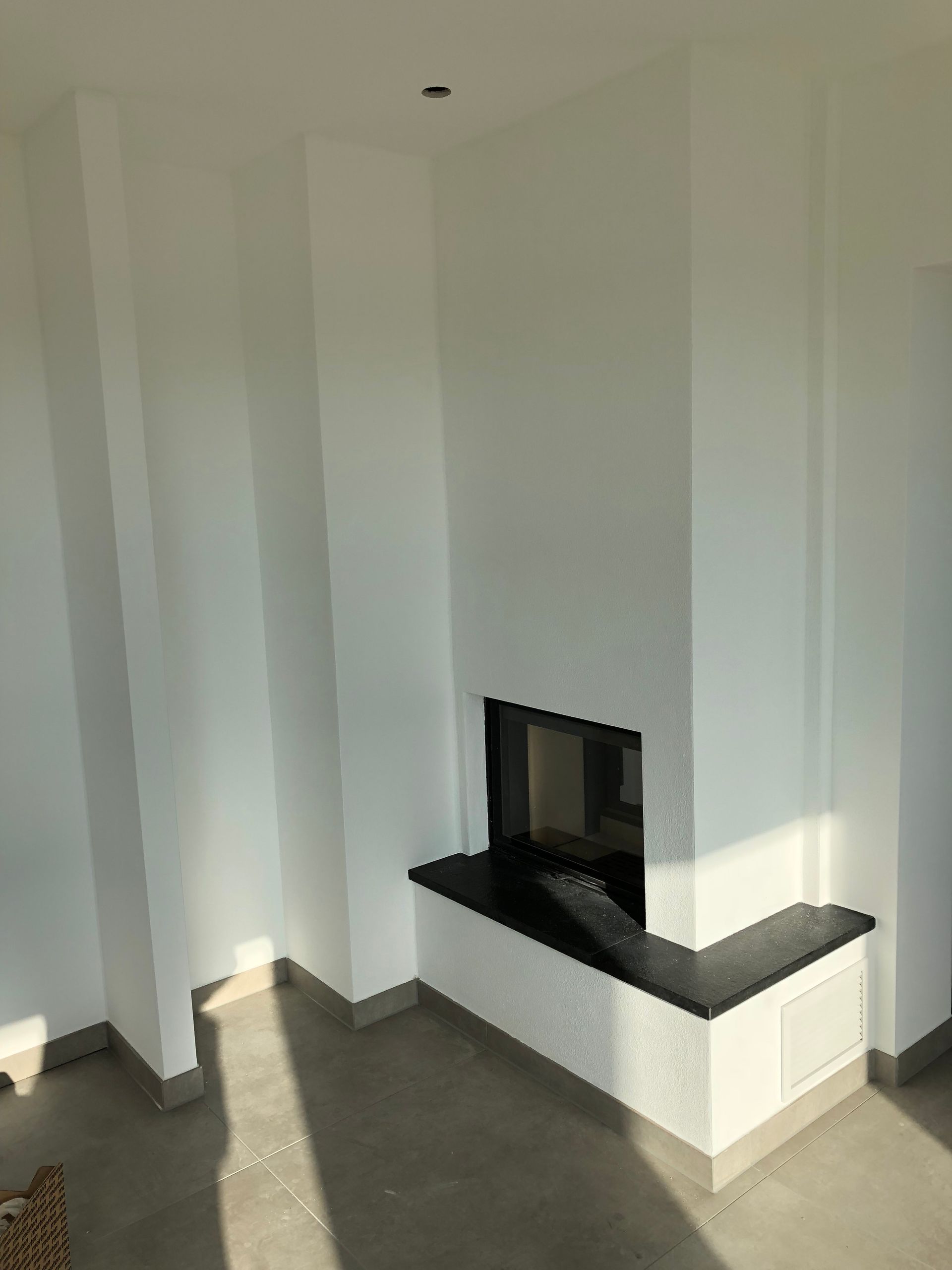 Weißer Eckkamin mit schwarzer Granitfeuerstelle in einem Raum mit weißen Wänden und grauem Boden.
