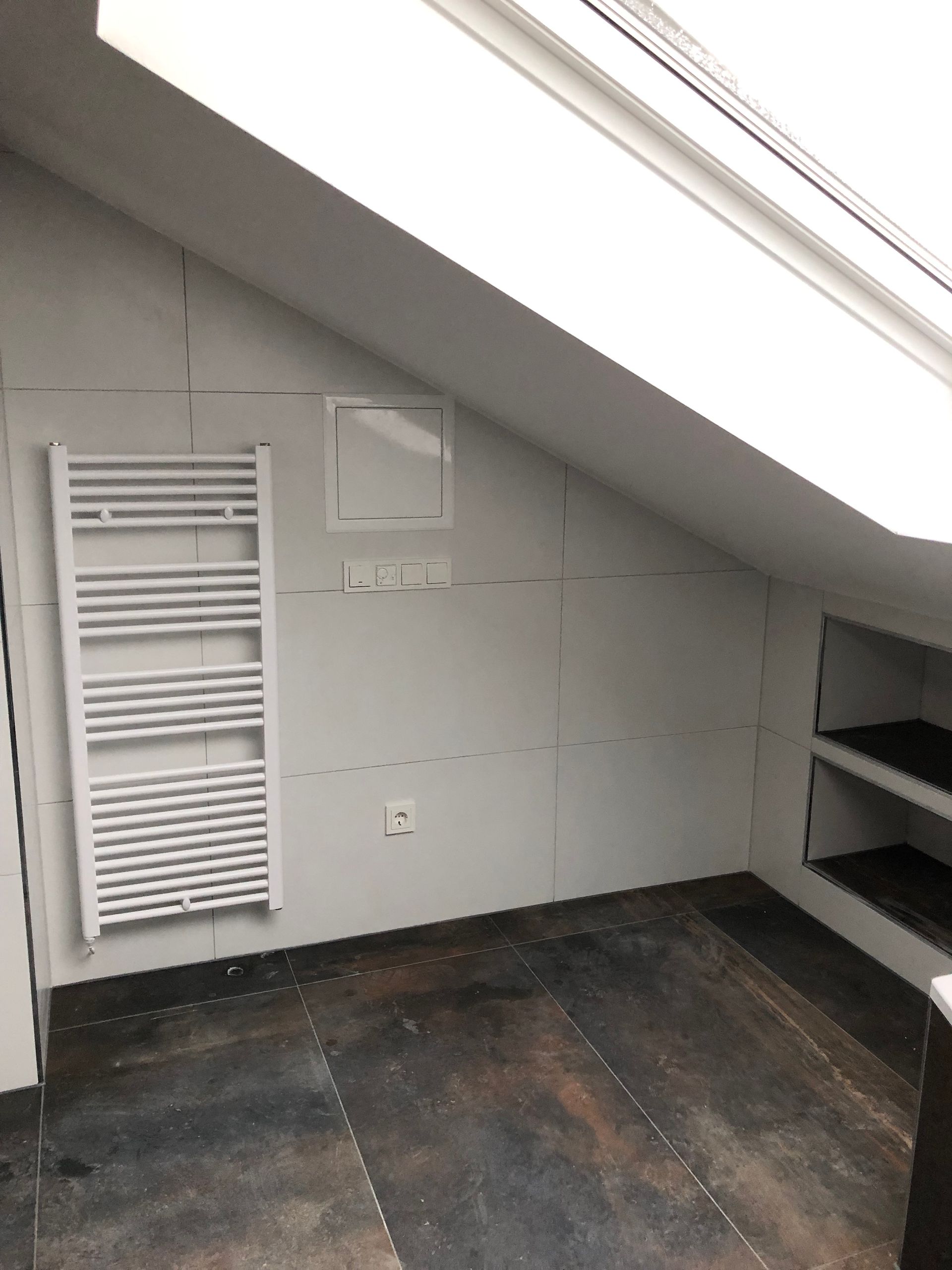 Weißer Heizkörper, Trockenbauwände und eingebaute Regale in einem schrägen Dachgeschossbad. Dunkler Holzboden.